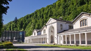 A large, white building with a classical architectural style, featuring columns and arched windows, surrounded by lush greenery and a clear blue sky.