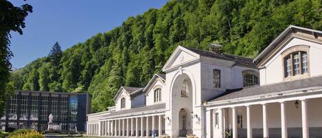 A large, white building with a classical architectural style, featuring columns and arched windows, surrounded by lush greenery and a clear blue sky.