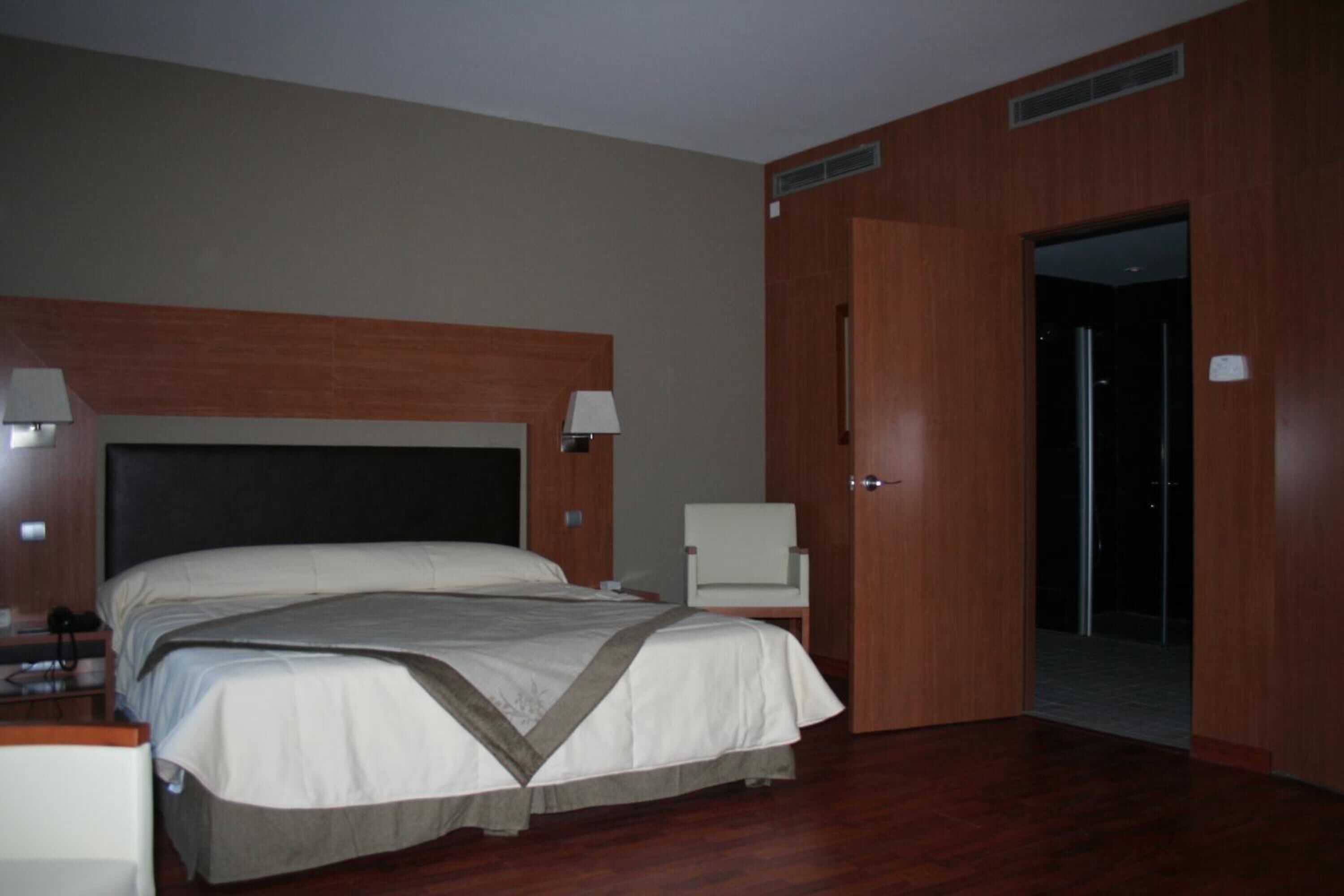 Ein Hotelzimmer mit einem großen Bett, Nachttischlampen, einem Sessel und einer Tür zu einem Flur.