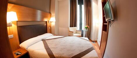 A hotel room with a large bed, bedside lamps, a TV mounted on the wall, and a window with curtains.