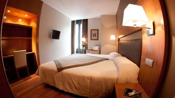 A hotel room with a bed, bedside table, desk, chair, TV, and wall-mounted lamps.