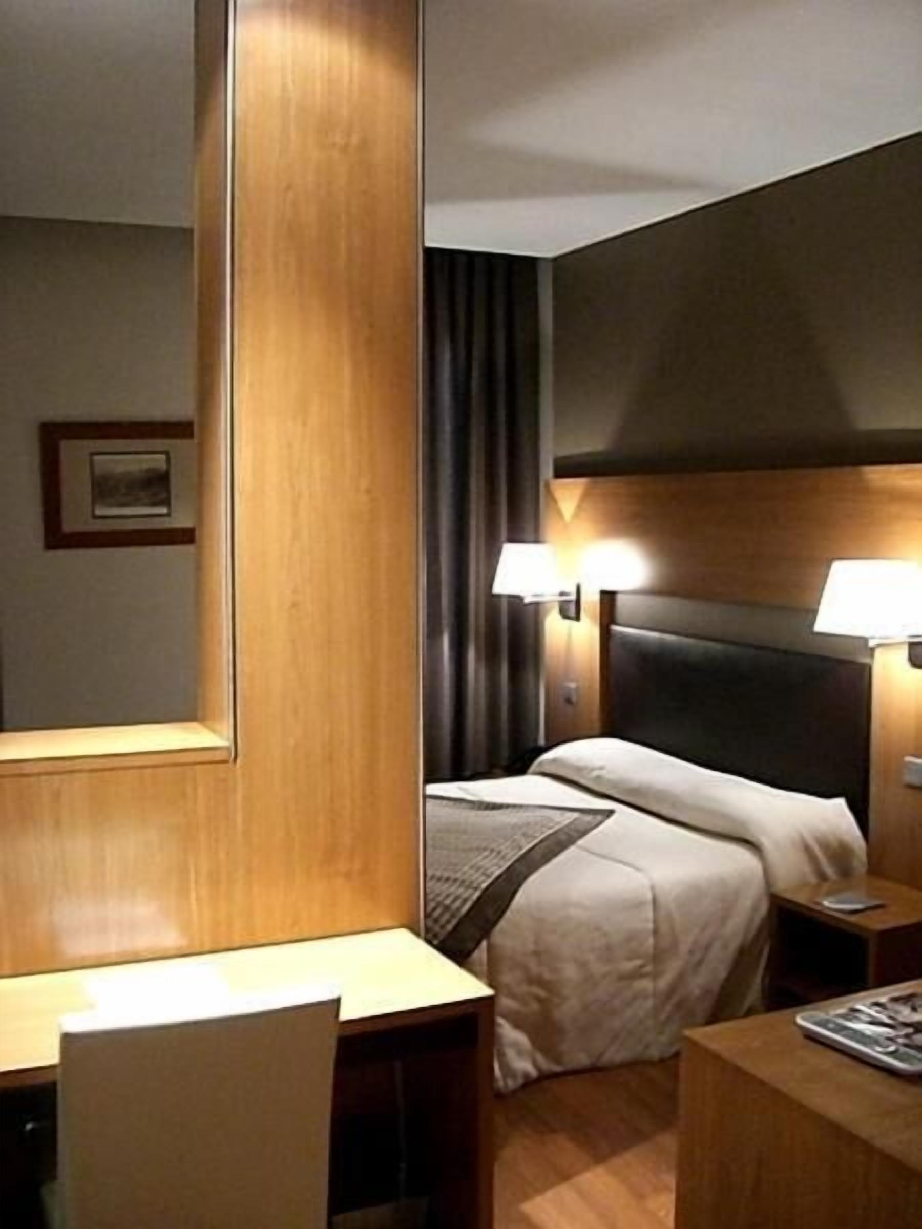 A hotel room with a wooden headboard, a bed with white linens, a nightstand with a lamp, and a desk with a chair.