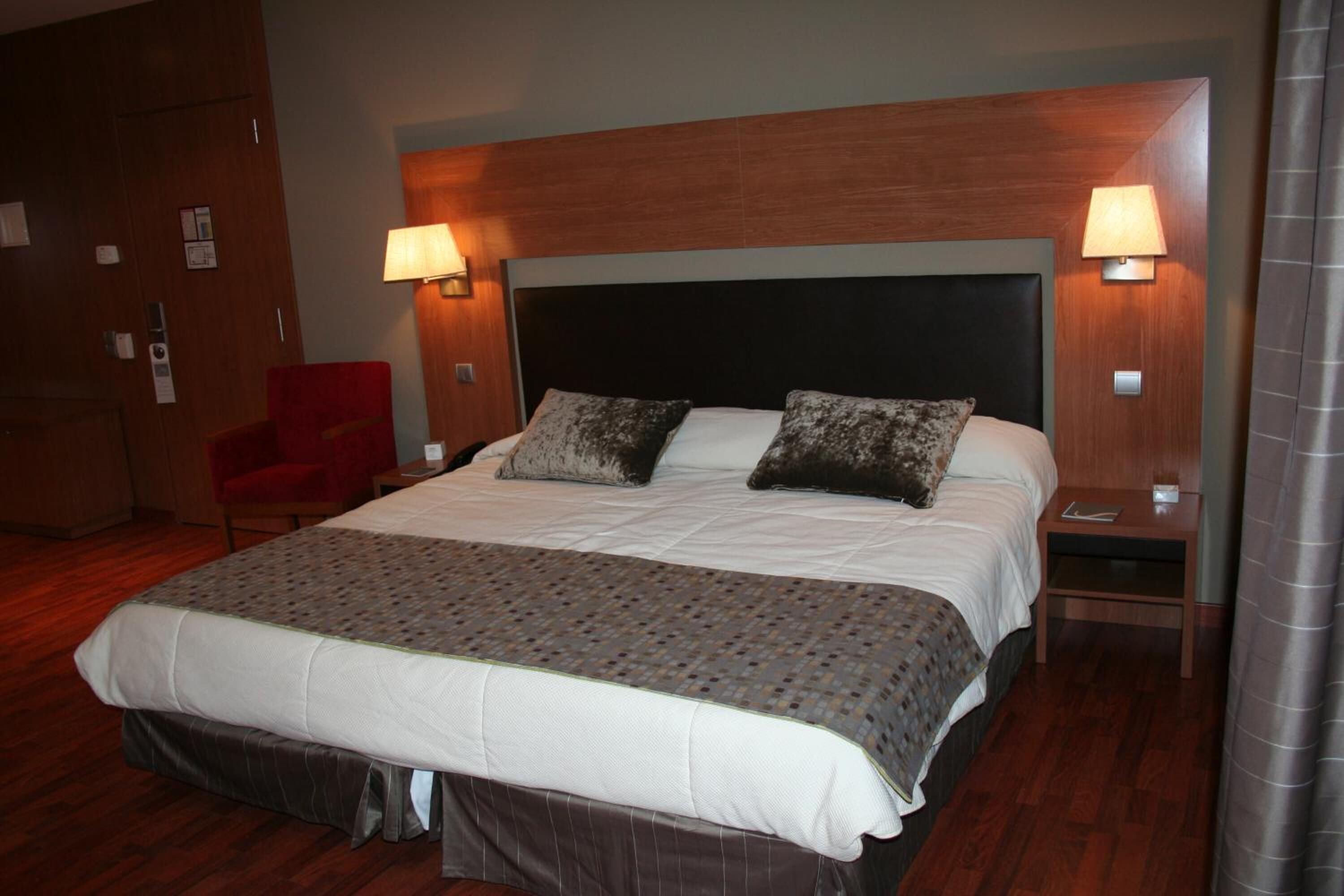 Habitación de hotel con una cama grande, dos lámparas de noche, una silla roja y un cabecero de madera.