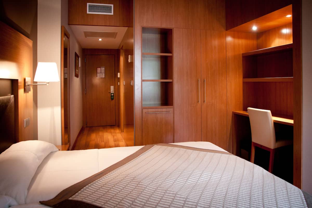 A hotel room with a bed, a desk, and a chair. There is a door leading to another room and a wall-mounted lamp.