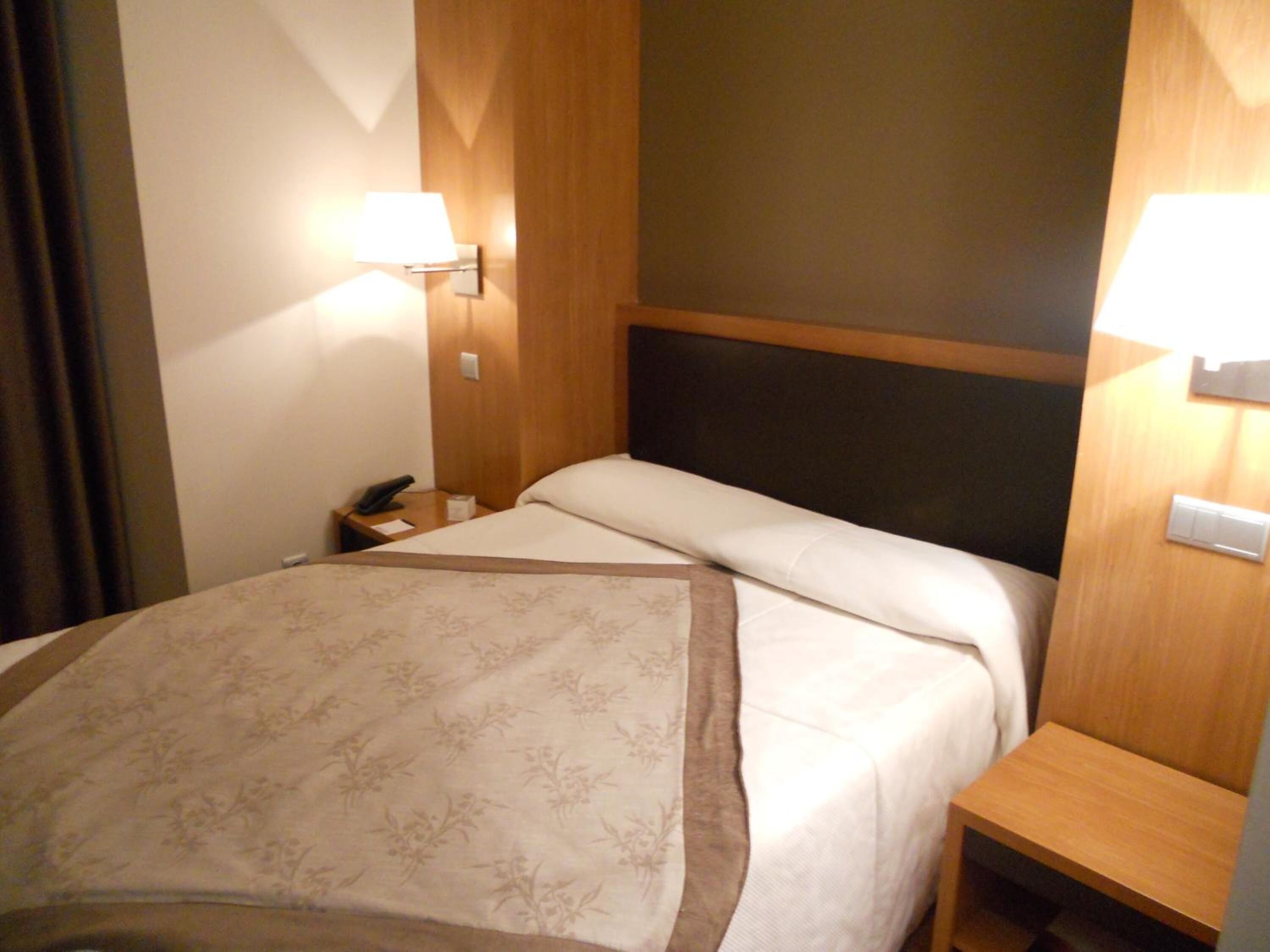 Una habitación de hotel con cama, mesitas de noche, lámparas y un cabecero de madera.