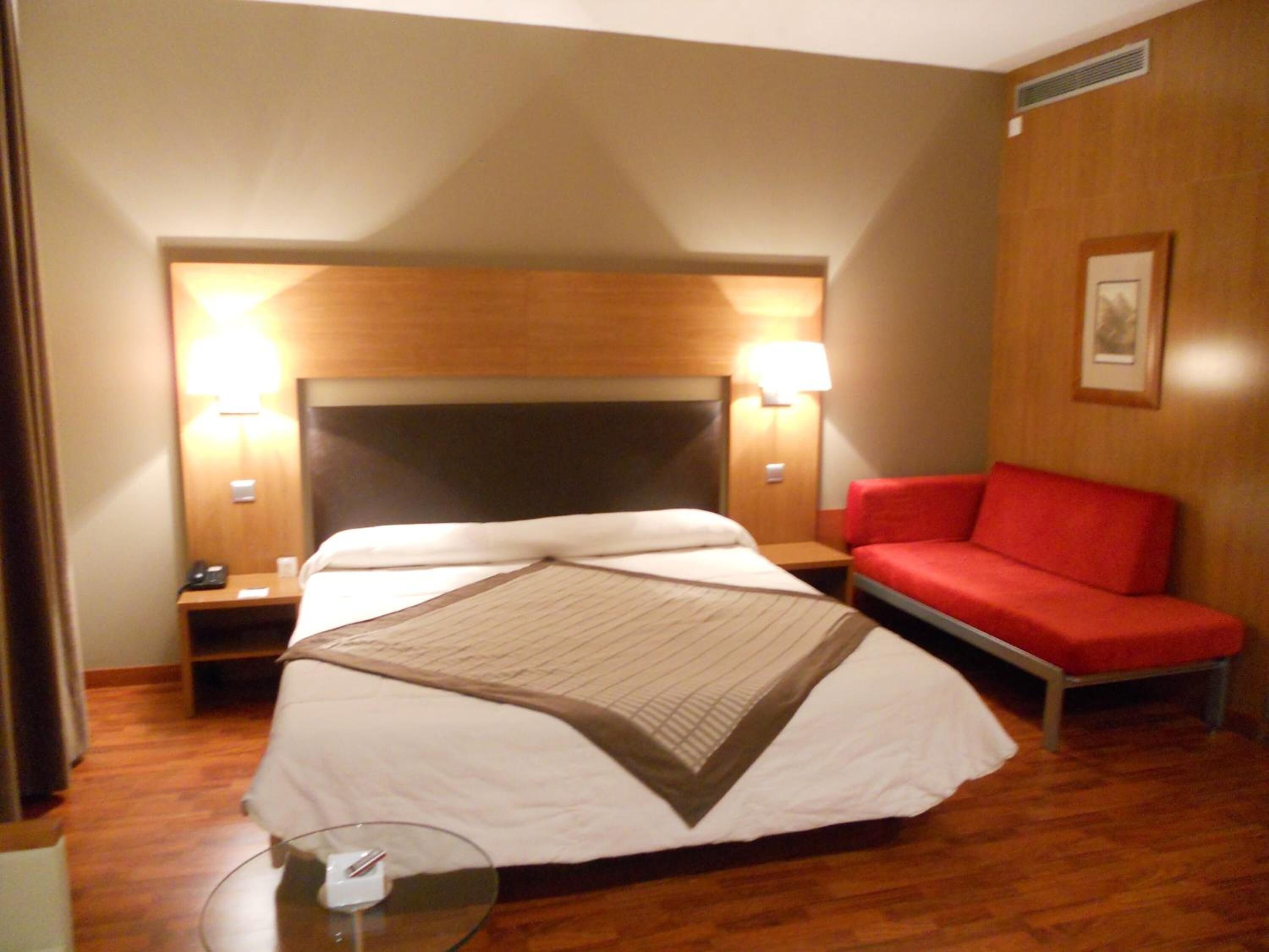 Una habitación de hotel con una cama grande, dos mesitas de noche, un sofá rojo y una pequeña mesa con superficie de vidrio.
