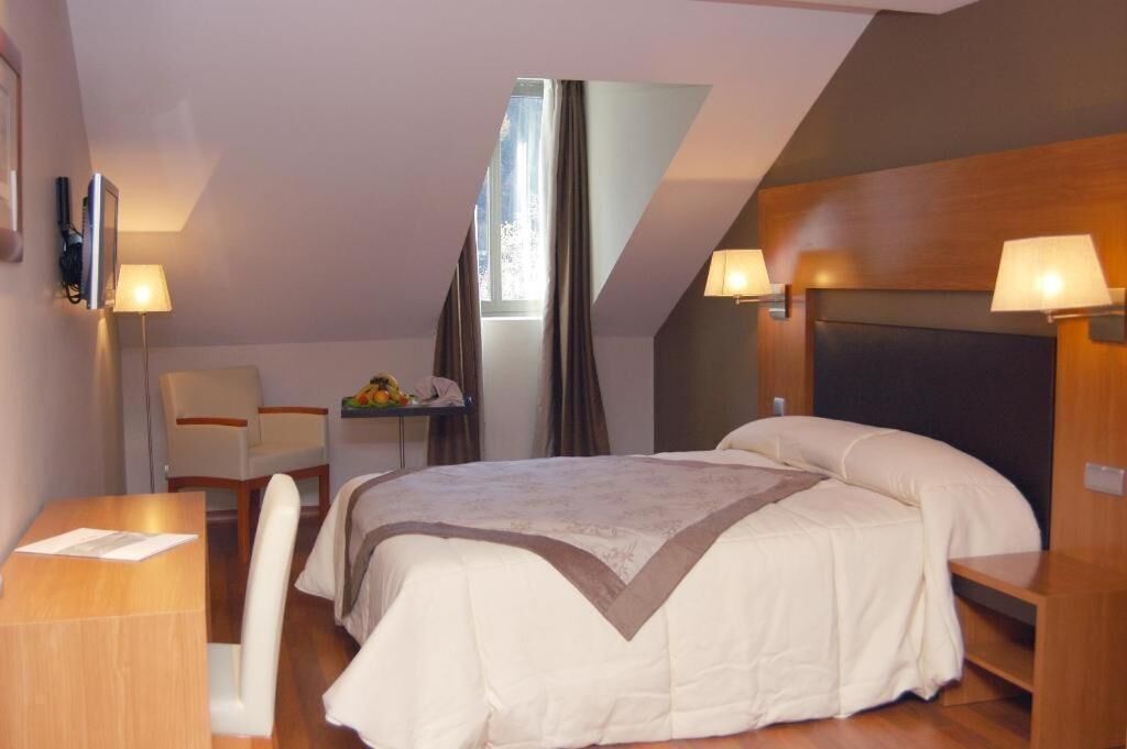 Un dormitorio con cama, mesitas de noche, escritorio y silla. Hay una ventana con cortinas, una lámpara en la pared y un televisor montado en la pared.