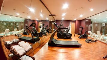 Une salle de sport équipée de plusieurs tapis de course, de vélos d’appartement et d’autres appareils d’exercice. On y trouve des chaises blanches et un miroir qui reflète la pièce.
