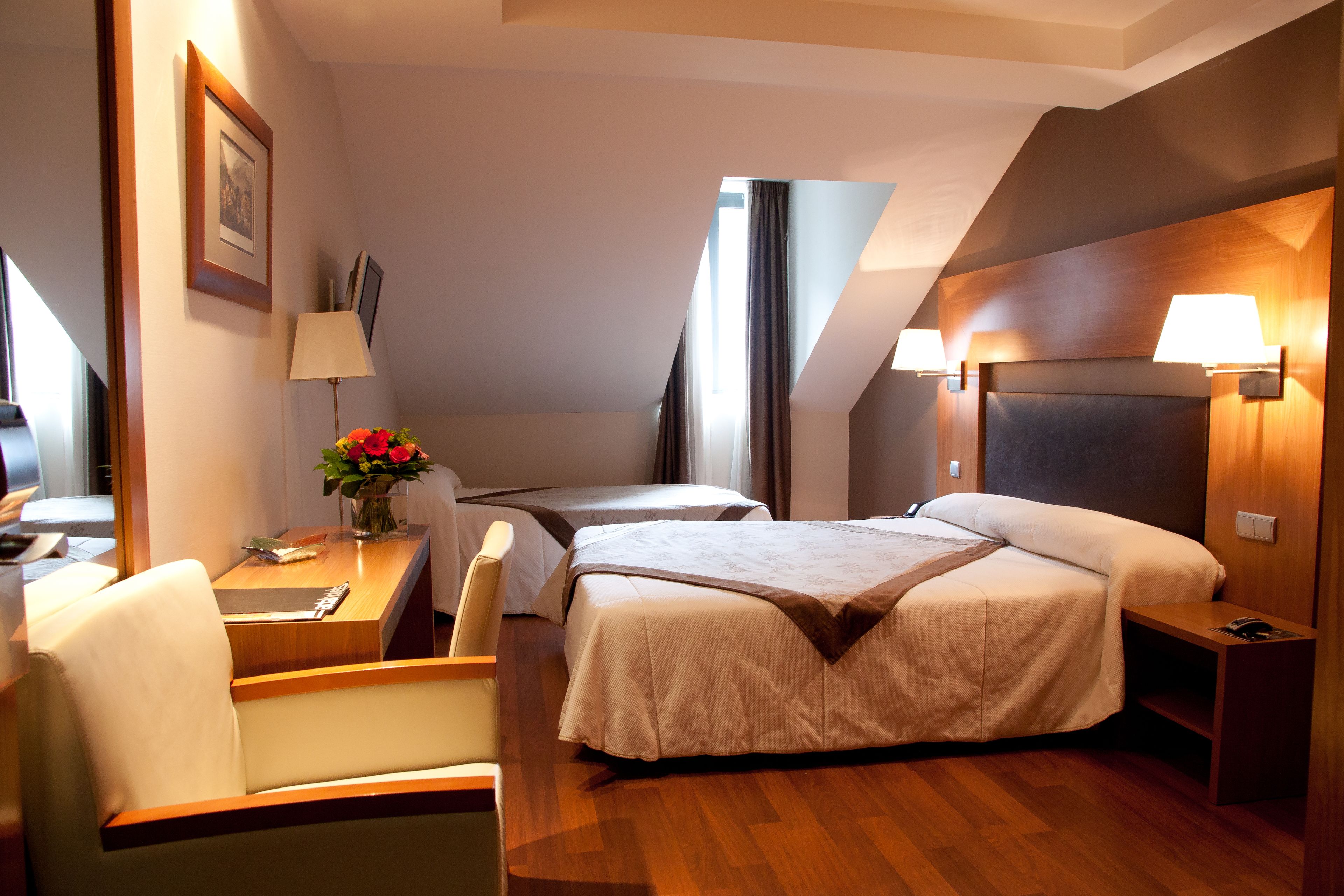 A hotel room with a large bed, a desk with a chair, a small table with a vase of flowers, and a lamp.