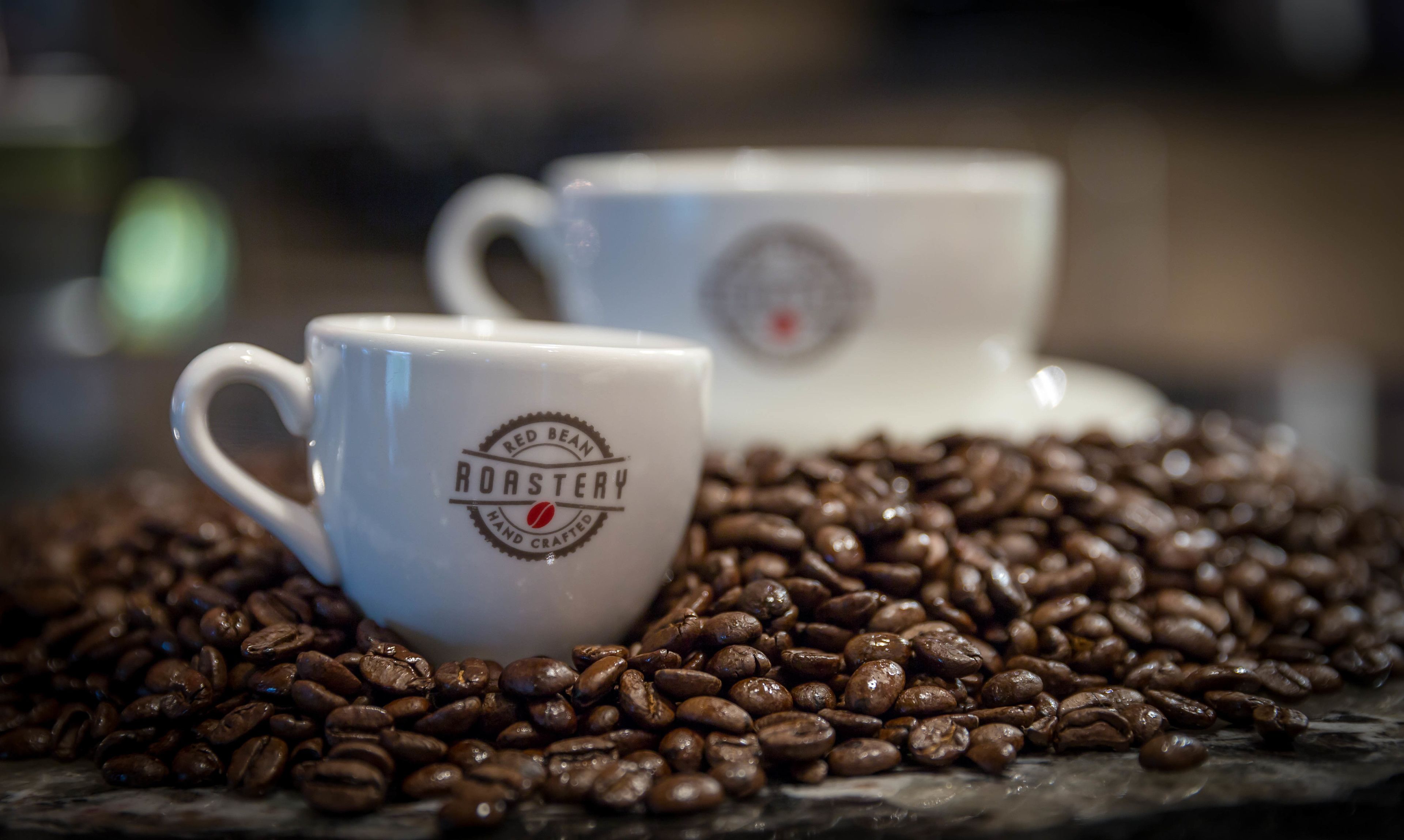 Two white ceramic cups with "Red Bean Roastery" branding, surrounded by coffee beans.