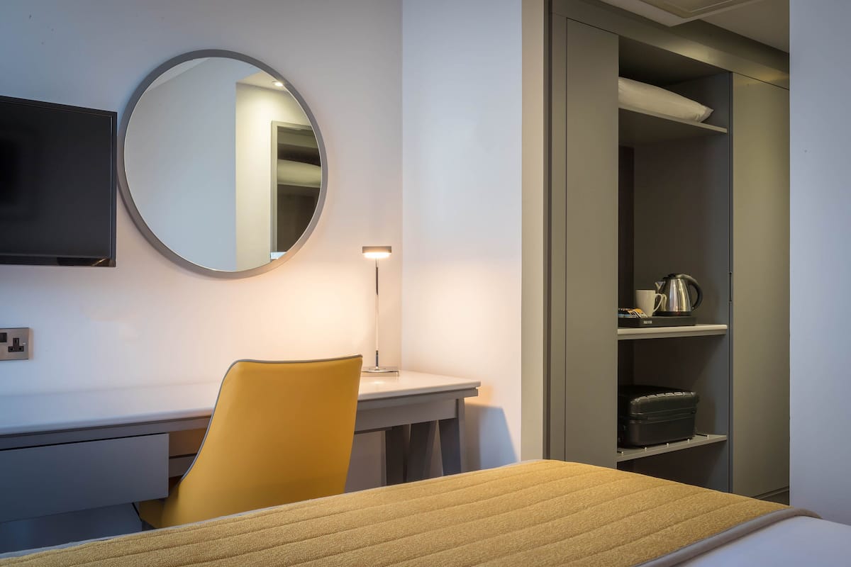Une chambre d’hôtel comprenant un lit, un bureau avec une chaise, un miroir rond, un téléviseur à écran plat, une lampe et une armoire avec des étagères.