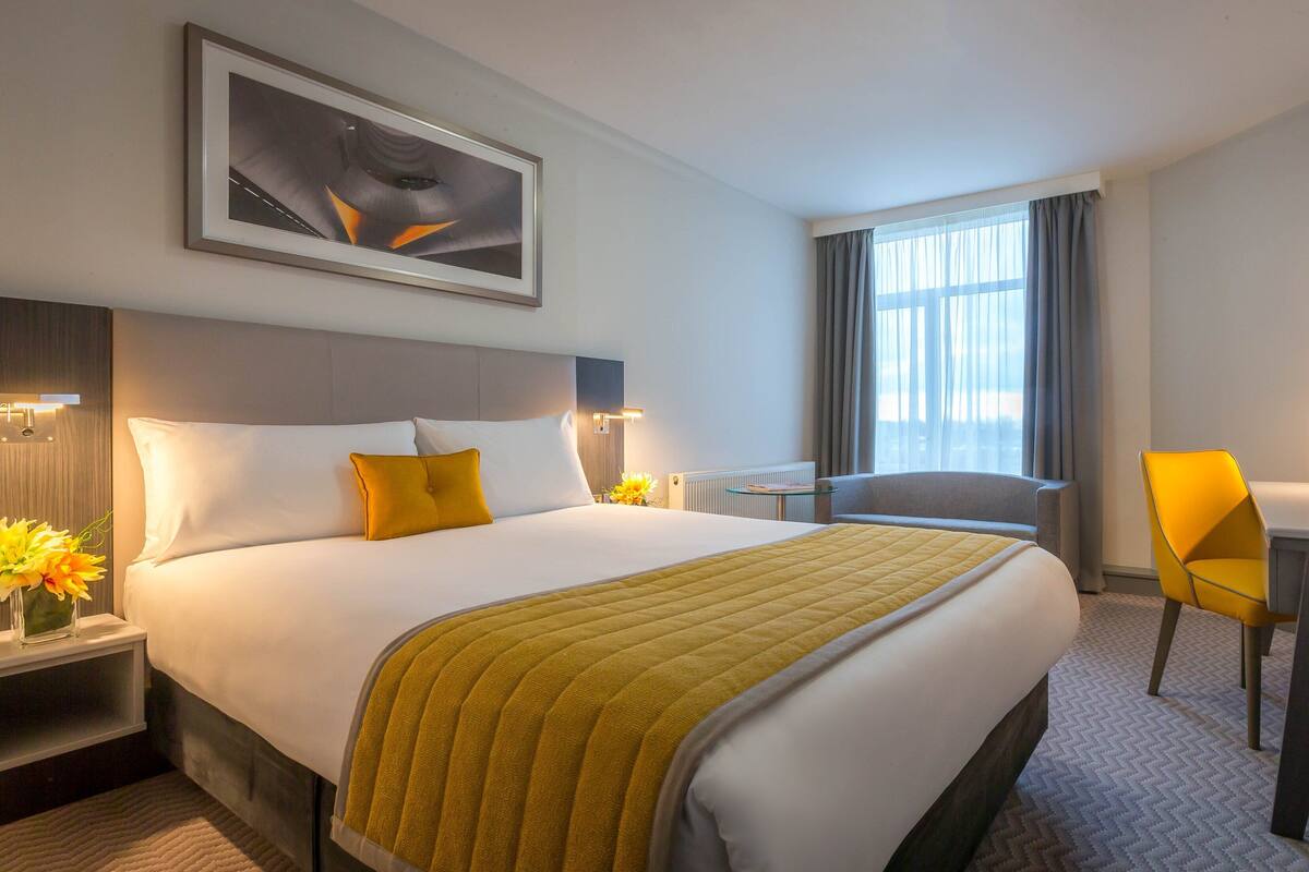 Une chambre d’hôtel avec un grand lit, un bureau, une chaise, une télévision et une fenêtre avec des rideaux.