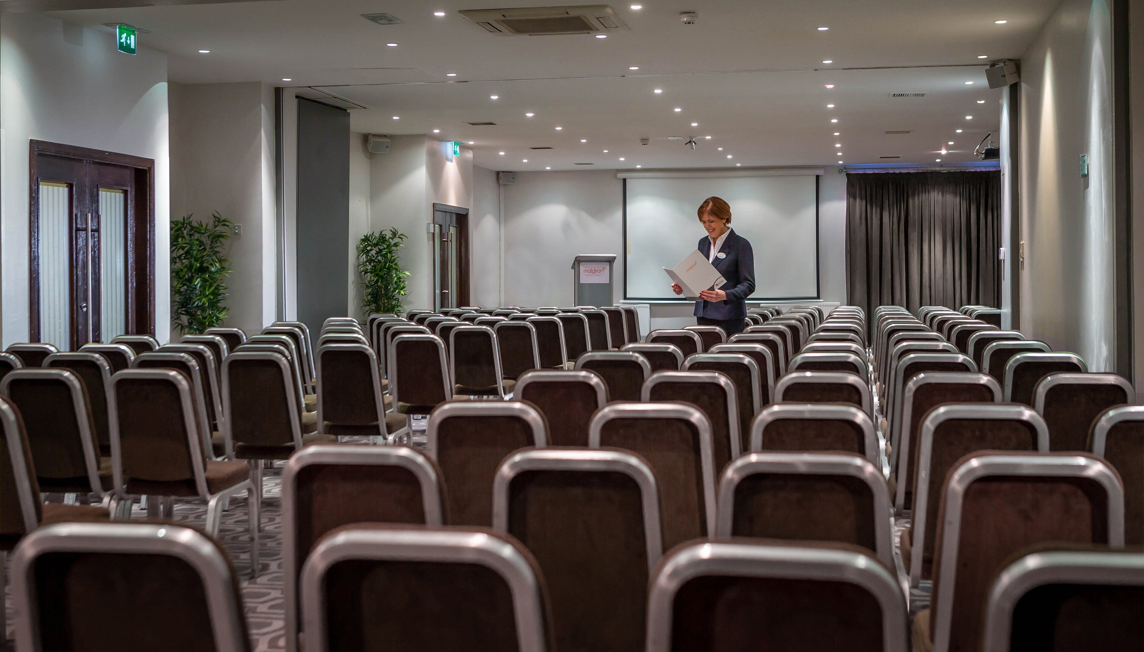 Un salón de conferencias con filas de sillas, un atril y una pantalla para presentaciones.