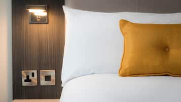 Habitación de hotel con cama blanca, almohada color mostaza, luz empotrada en la pared y dos enchufes.