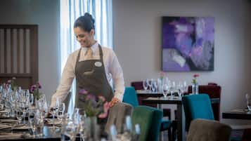 Un restaurante con un camarero uniformado, mesas dispuestas con copas de vino y platos, y un cuadro abstracto moderno en la pared.