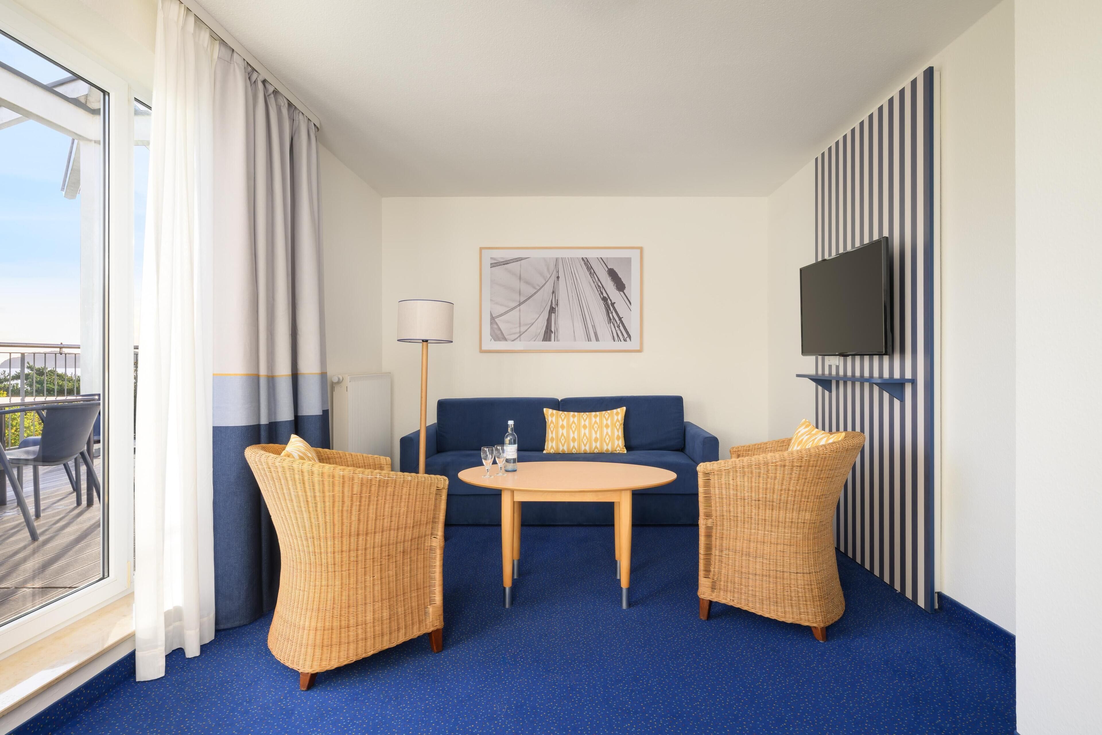 A hotel room with a blue carpet, a sofa, two wicker chairs, a small round table, a lamp, a TV mounted on a striped wall, and a window with curtains.