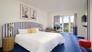 Habitación de hotel con una cama grande, una mesita pequeña, un sofá, un televisor y un balcón con cortinas.