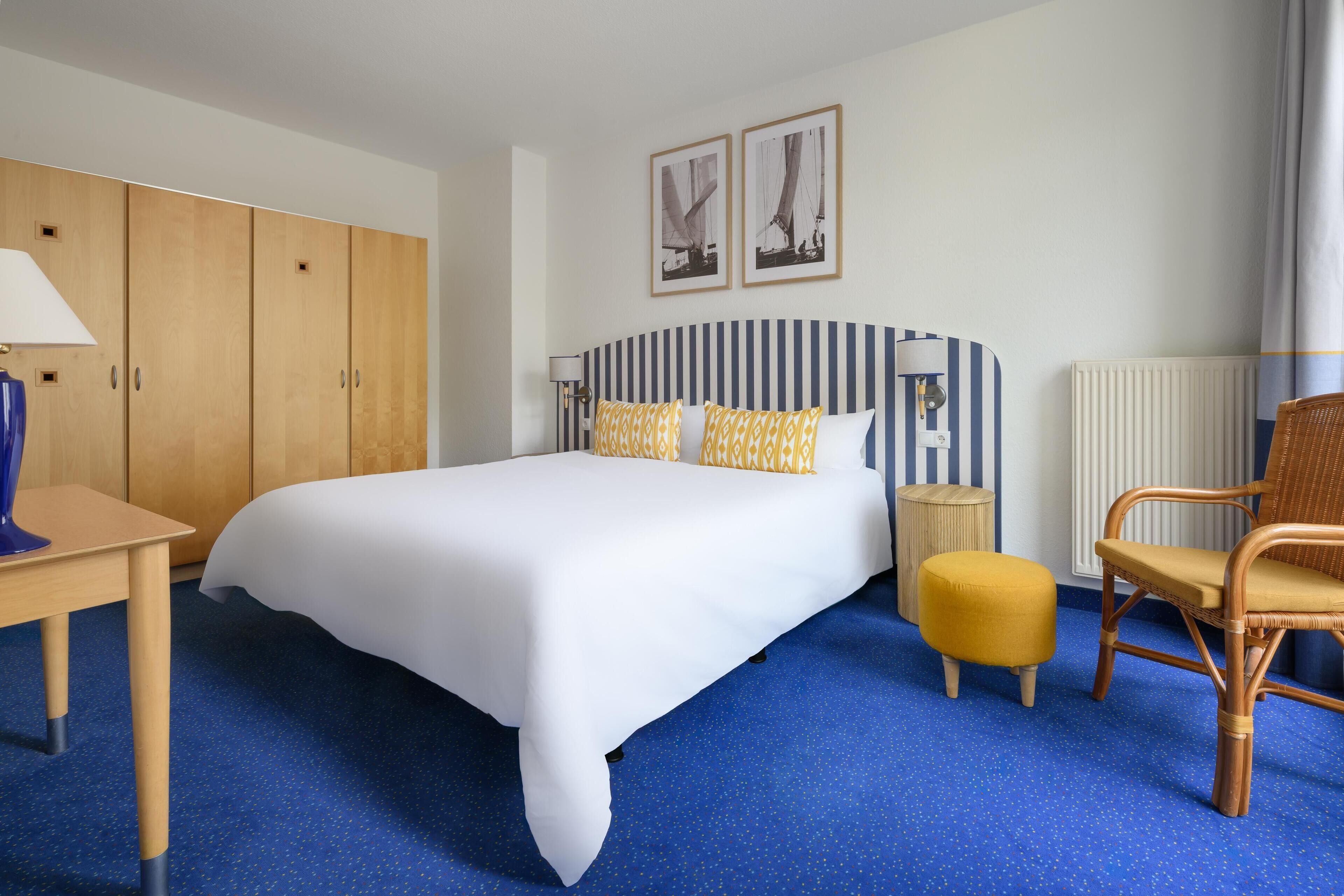 Une chambre d’hôtel avec un grand lit, une armoire en bois, une tête de lit à rayures, une chaise en osier et une petite table ronde.