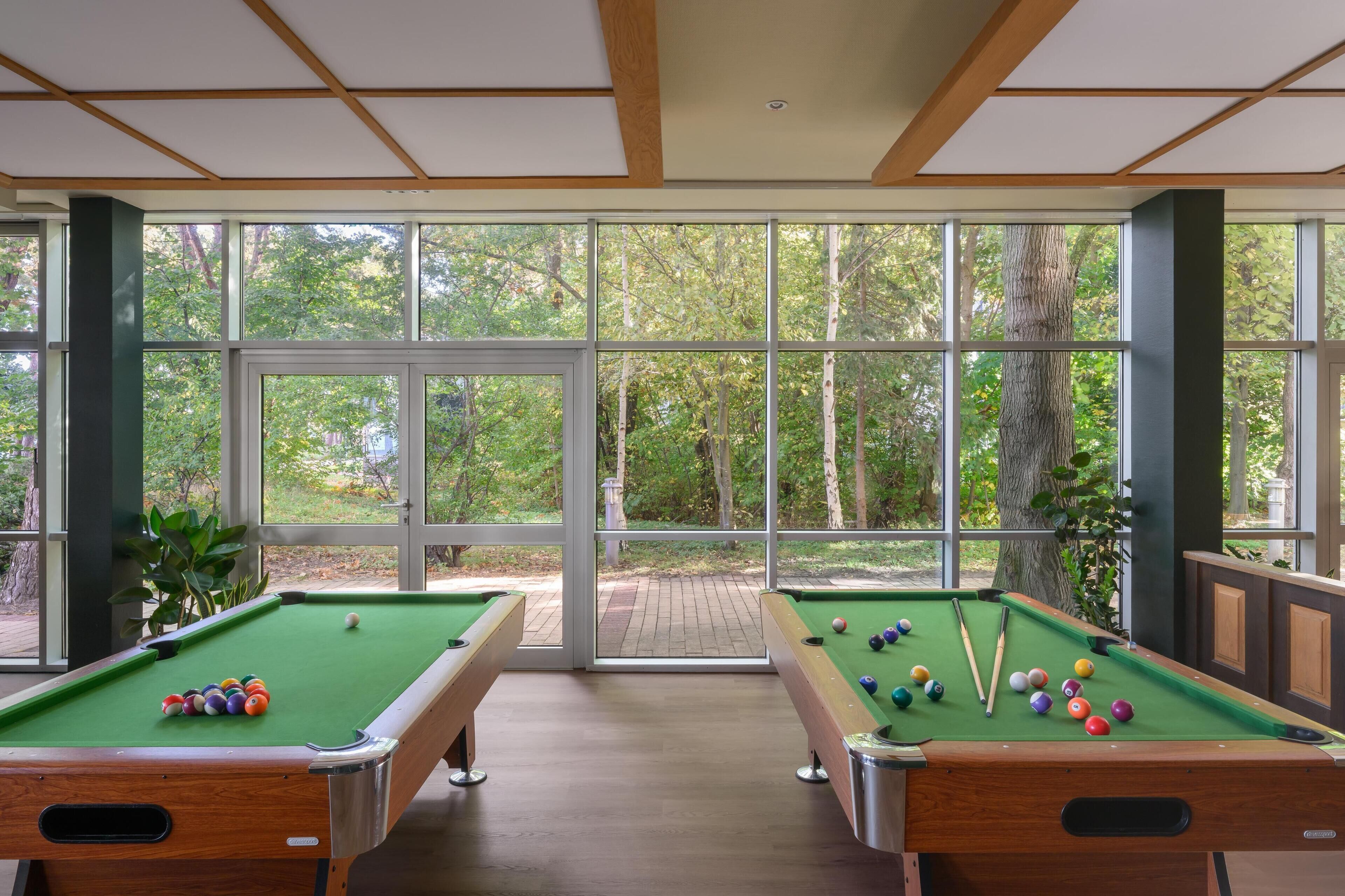 A pool table with balls and cues, large windows with a view of trees, and a wooden ceiling.