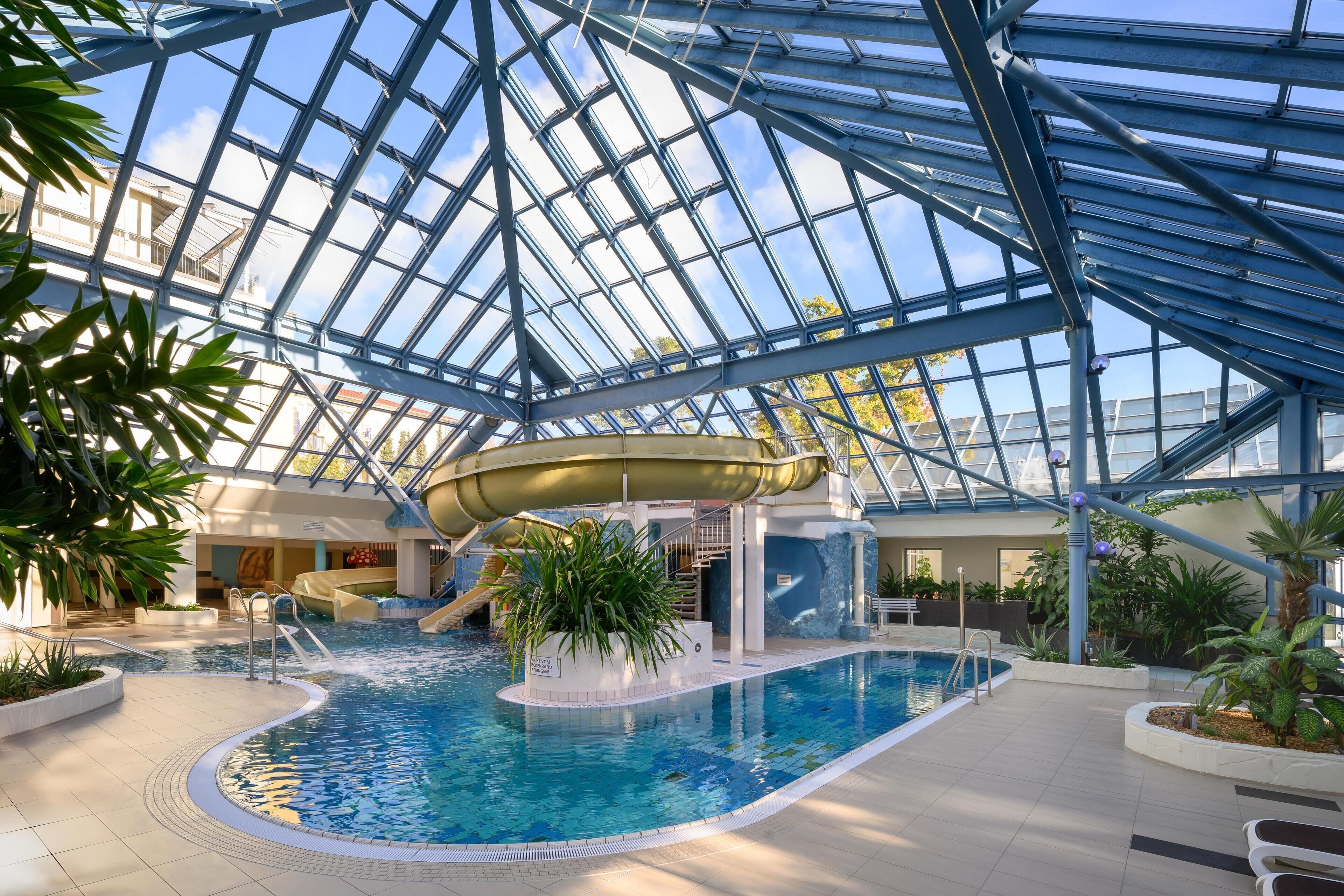 An indoor swimming pool with a glass ceiling, surrounded by plants and a slide.