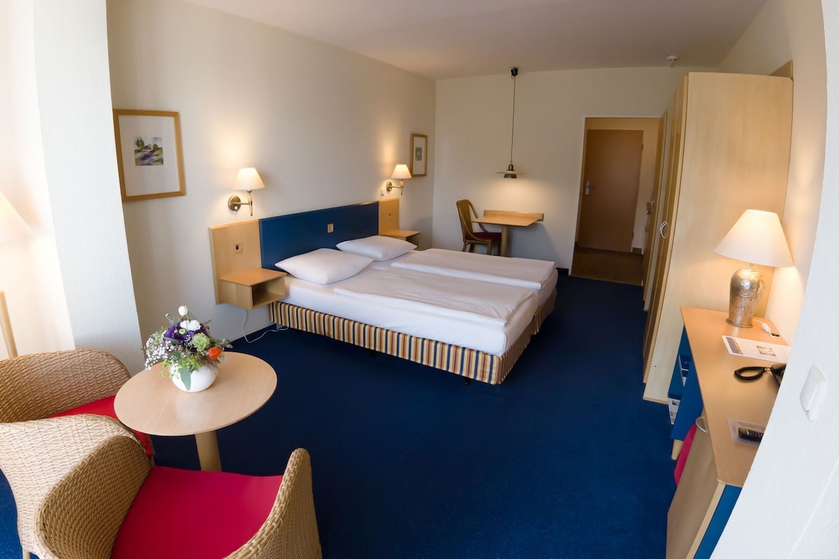 A hotel room with a bed, two bedside tables, a chair, a small table with a vase, and a door.