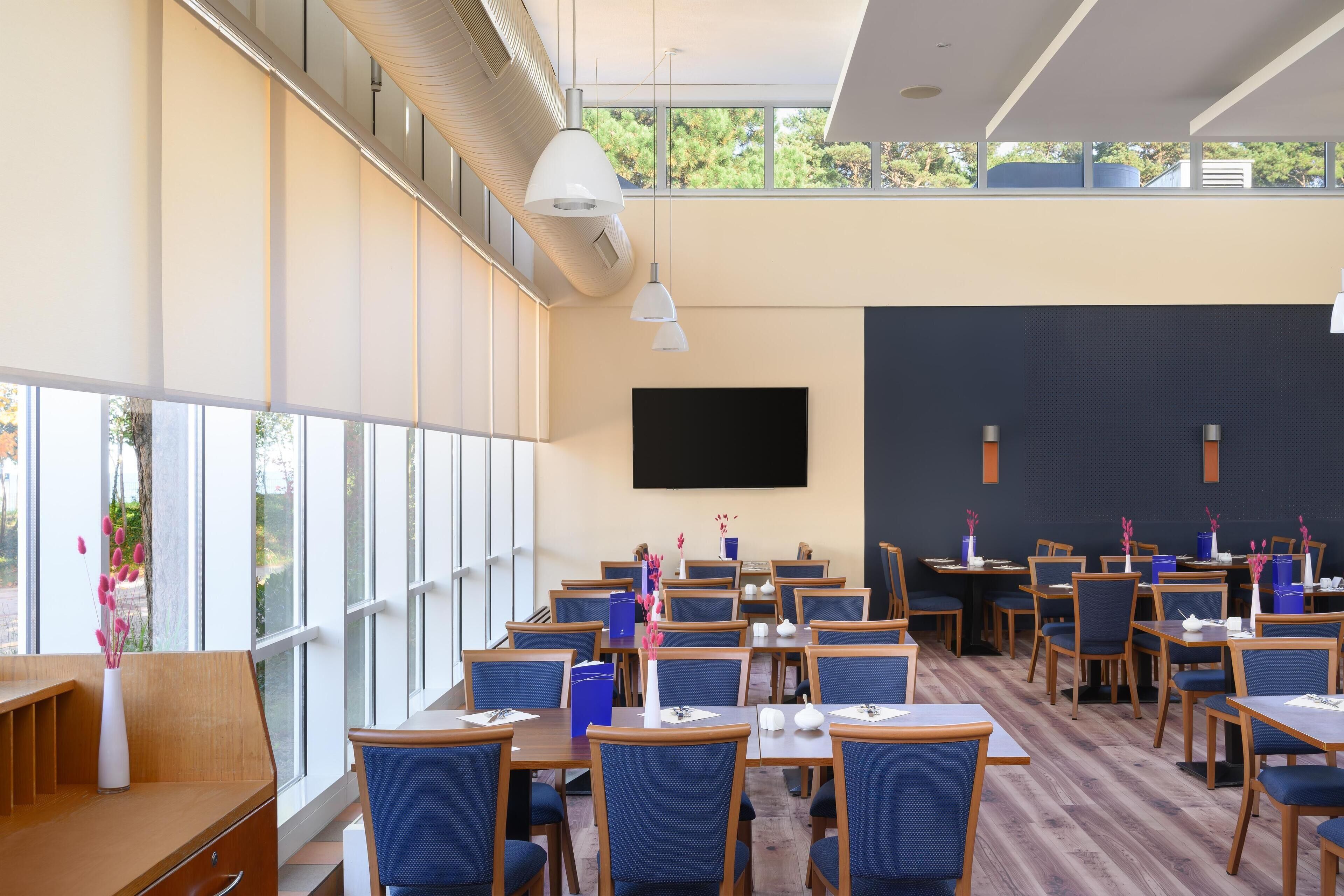 A modern dining area with multiple tables and chairs, large windows, and a flat-screen TV on the wall.