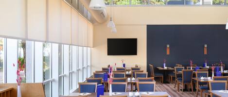 A modern dining area with multiple tables and chairs, large windows, and a flat-screen TV on the wall.