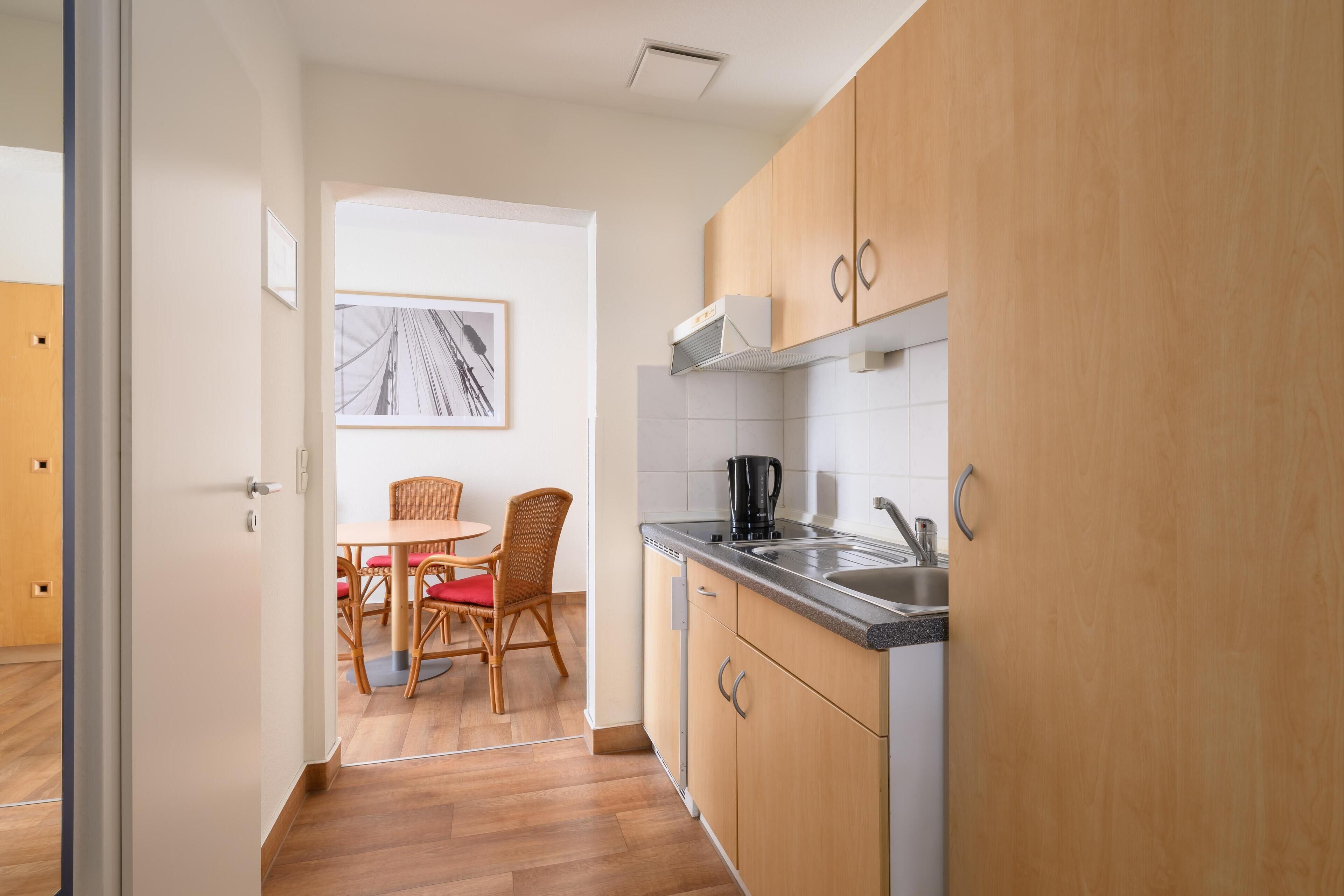 A modern kitchen with wooden cabinets, a stainless steel sink, and a dining area with wicker chairs and a red cushion.