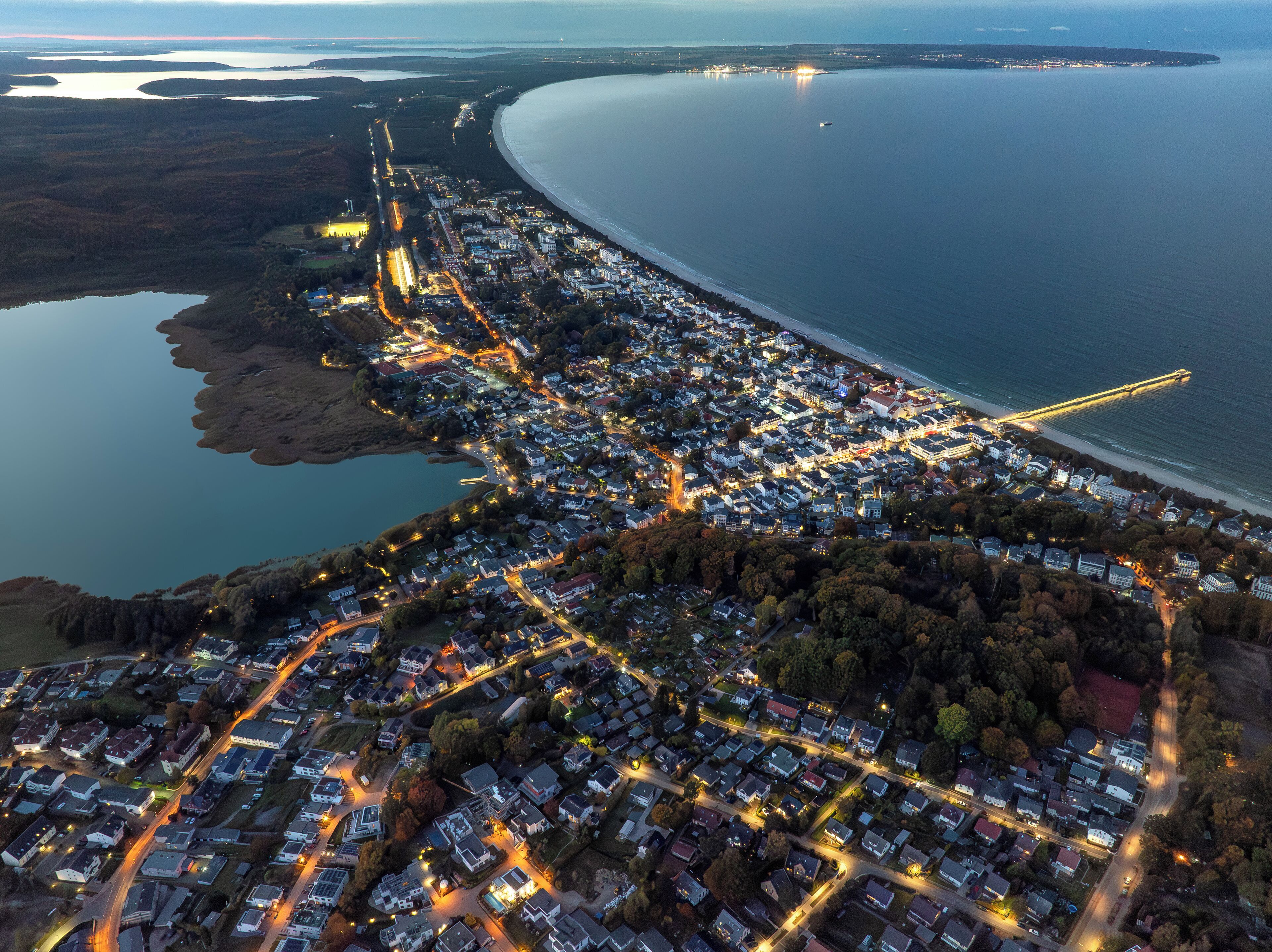An aerial view of a coastal town with a long beachfront, well-lit streets, and a bridge connecting the mainland to an island.