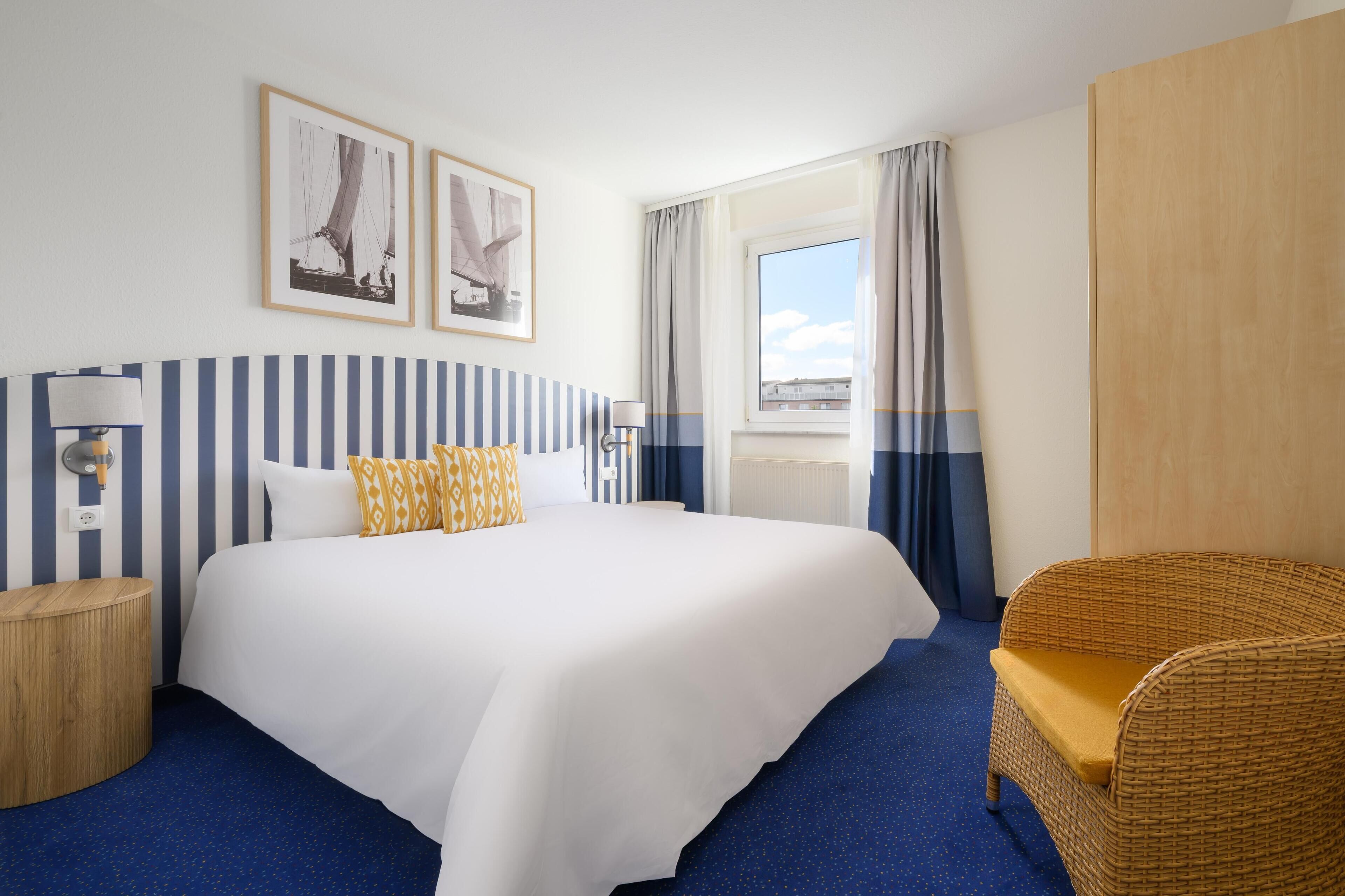 A hotel room with a large bed, a wicker chair, a window with curtains, and two framed pictures on the wall.