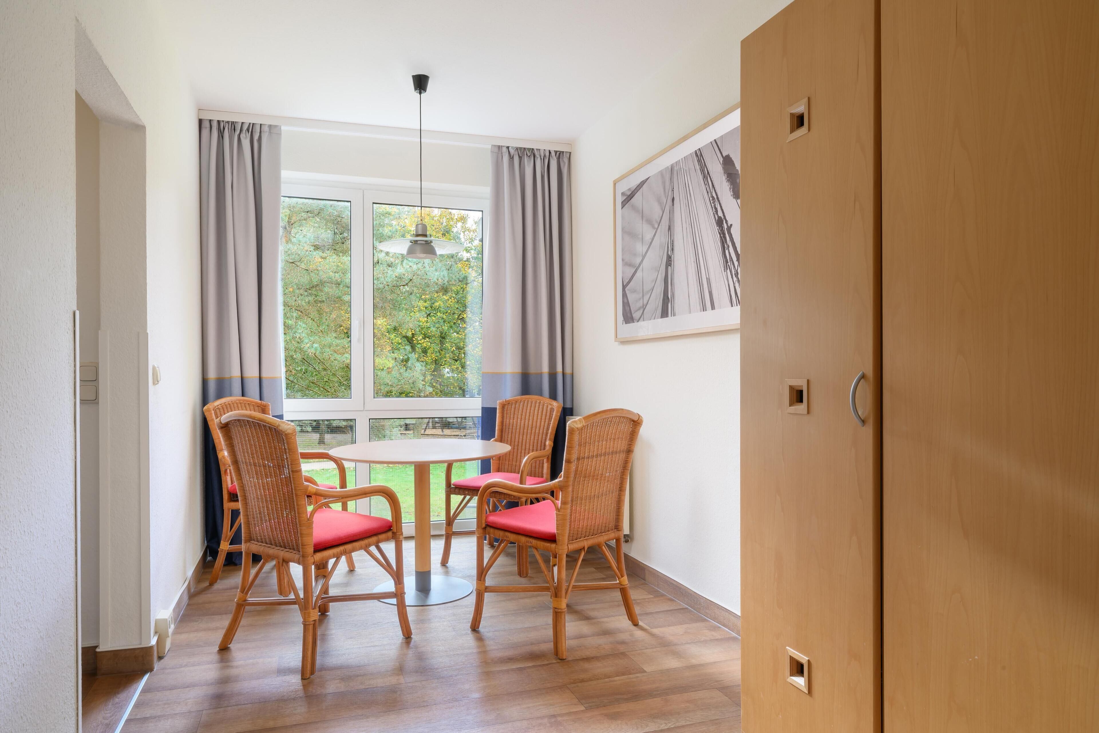 A room with a round table and four wicker chairs with red cushions, a window with curtains, and a framed picture on the wall.