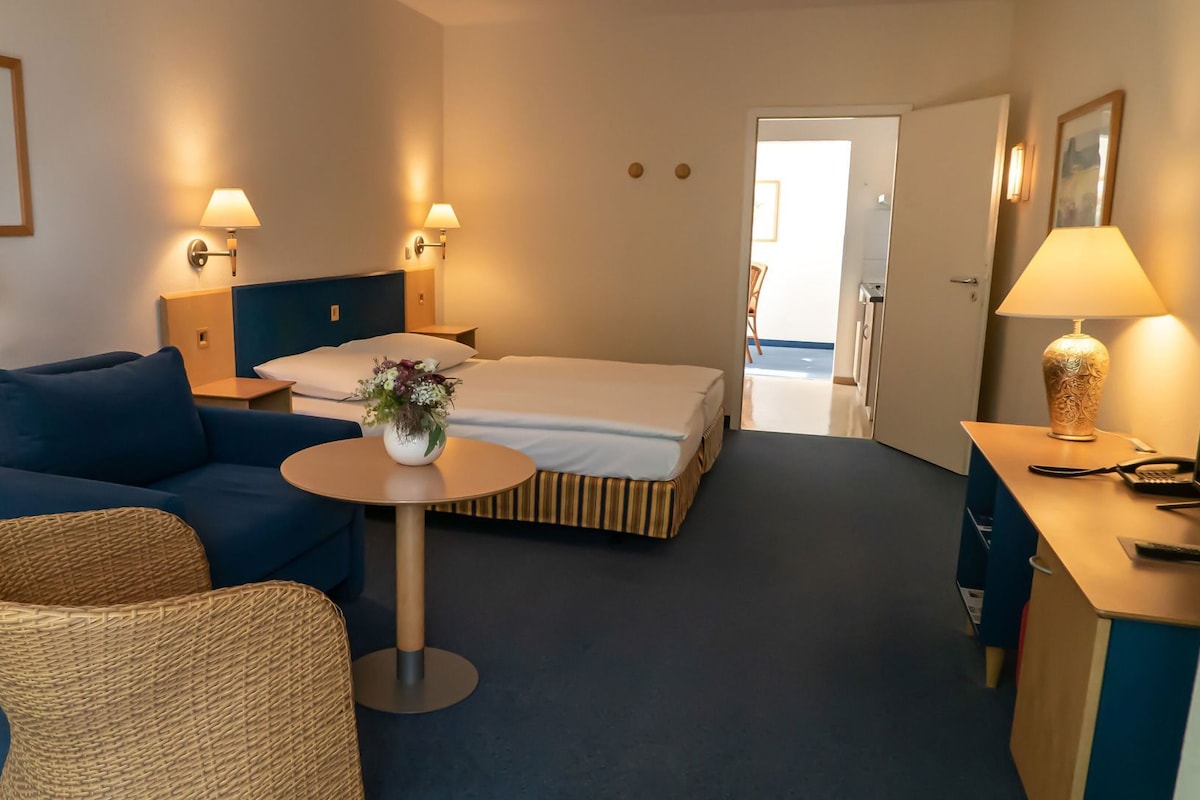 A hotel room with a bed, a sofa, a small table with a vase, and a desk with a lamp.