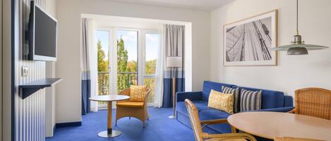 Habitación de hotel con alfombra azul, sofá con cojines estampados, mesa de comedor con sillas, televisor montado en la pared y balcón con cortinas.