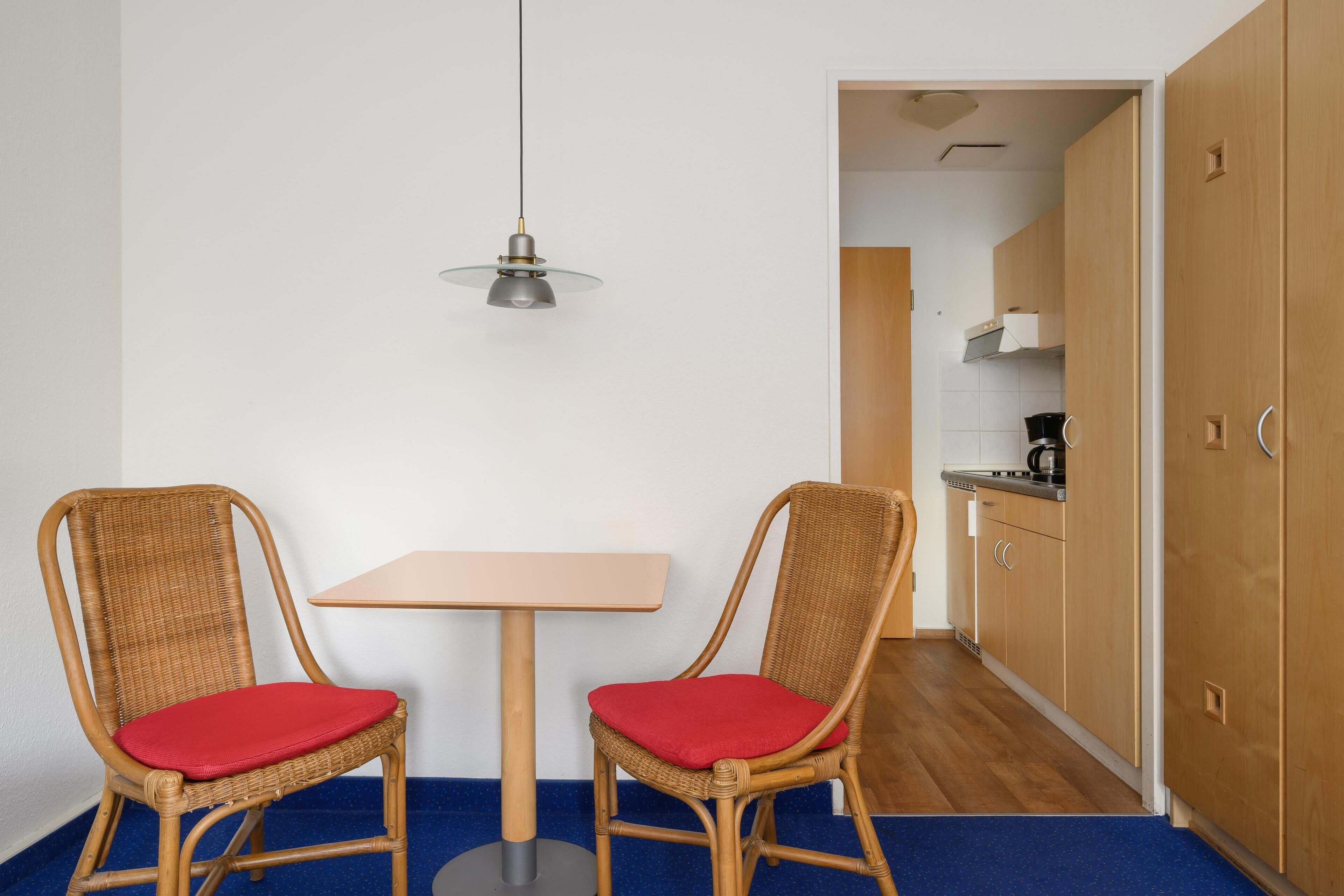 Una habitación pequeña con una mesa, dos sillas, una lámpara colgante y armarios de madera.