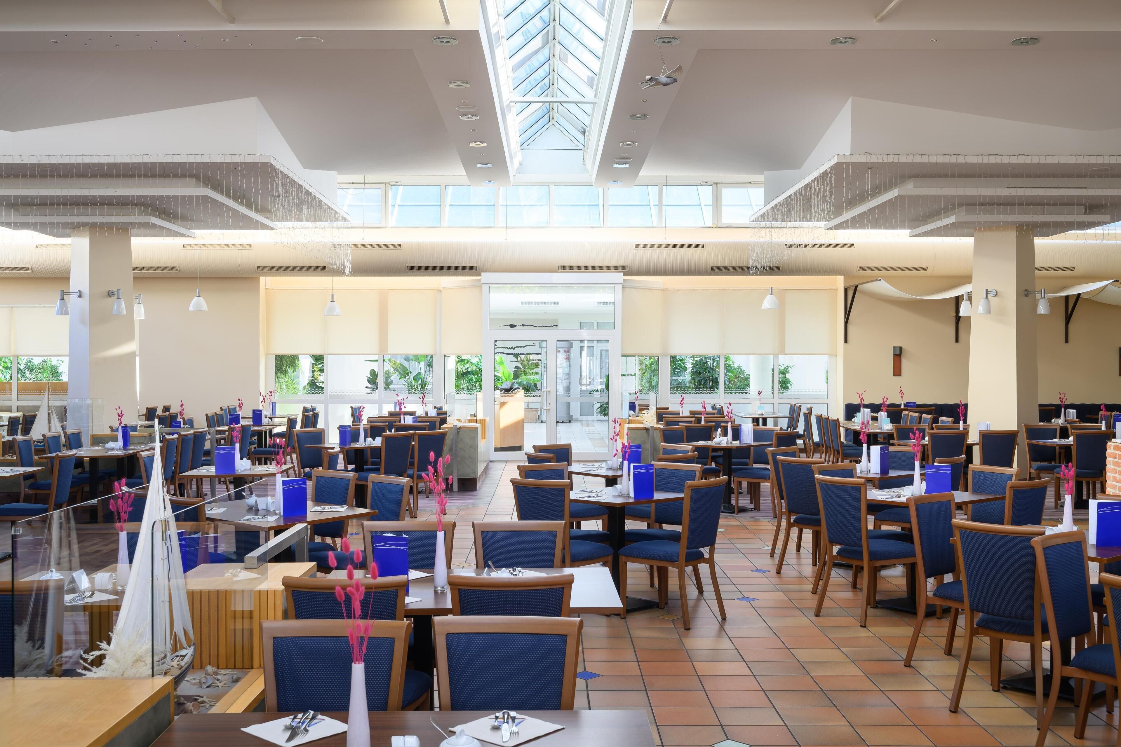 A spacious indoor dining area with multiple tables and chairs, large windows allowing natural light, and a central open space with a view of the outdoors.
