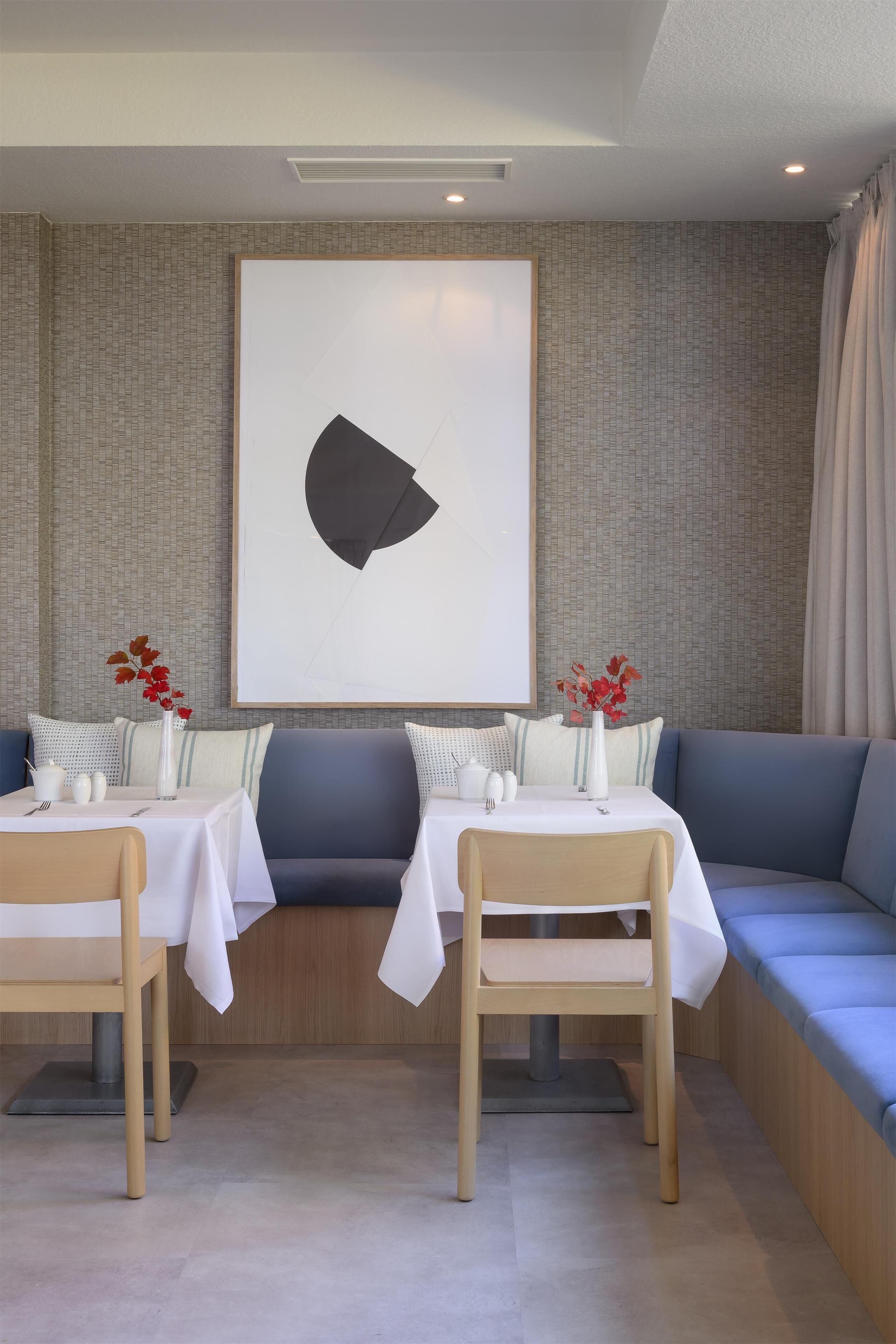 A modern dining area with a large abstract painting, two tables with white tablecloths, blue cushioned seating, and red flowers in vases.