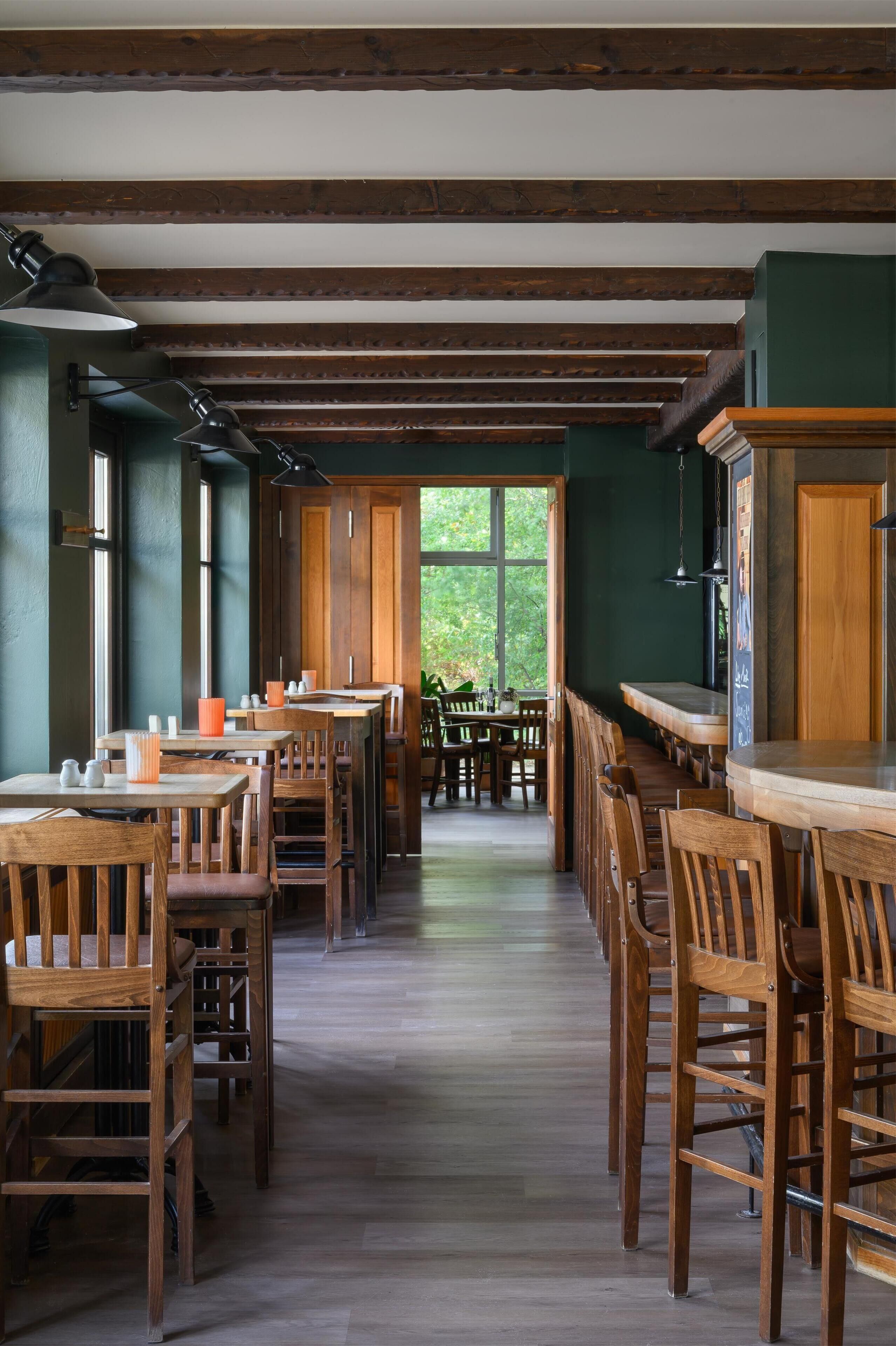 Une grande salle à manger avec des tables et des chaises en bois, un plafond haut avec des poutres apparentes et de grandes fenêtres laissant entrer la lumière naturelle.