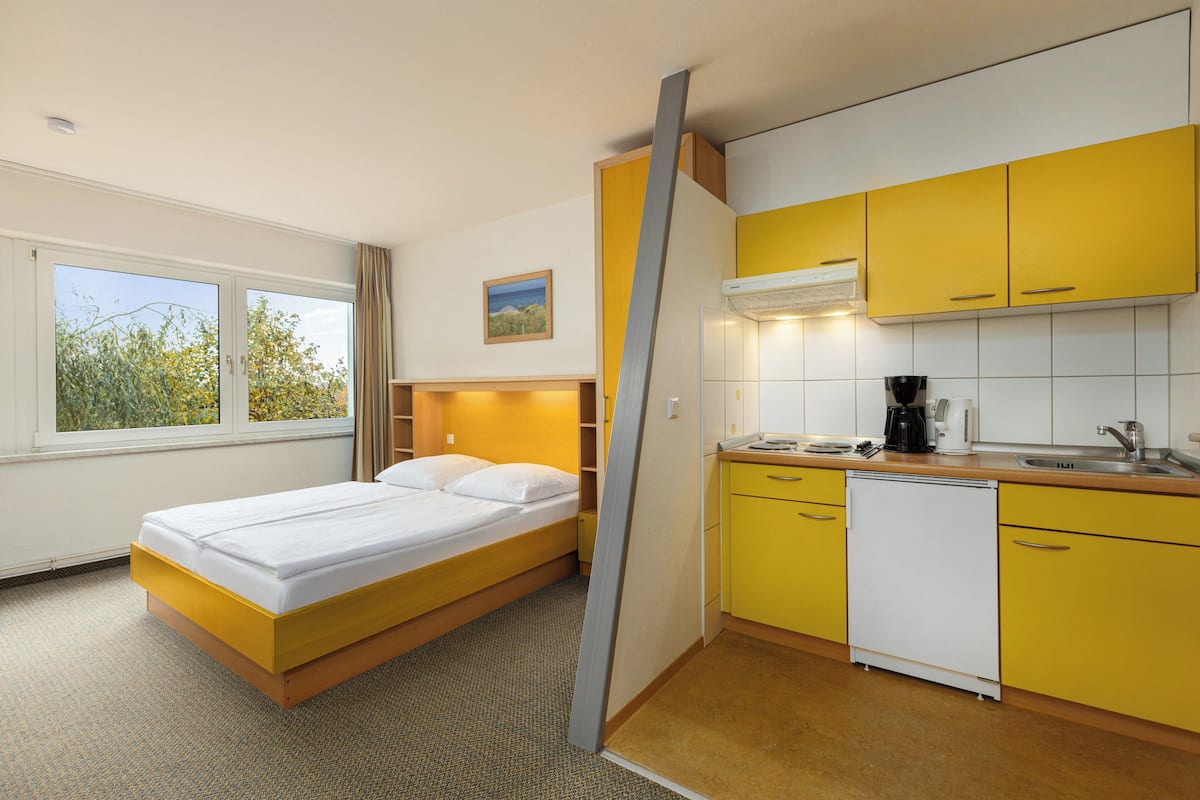 Una habitación de hotel compacta con una cama, un pequeño rincón de cocina y una ventana con vistas a árboles.