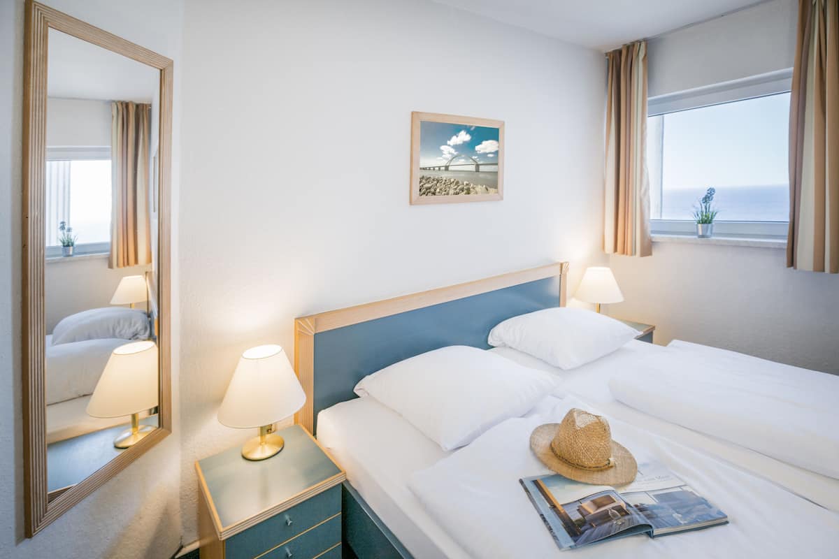 Una habitación de hotel con una cama, dos lámparas de noche, una mesita de noche, un espejo, un cuadro en la pared y una ventana con cortinas.