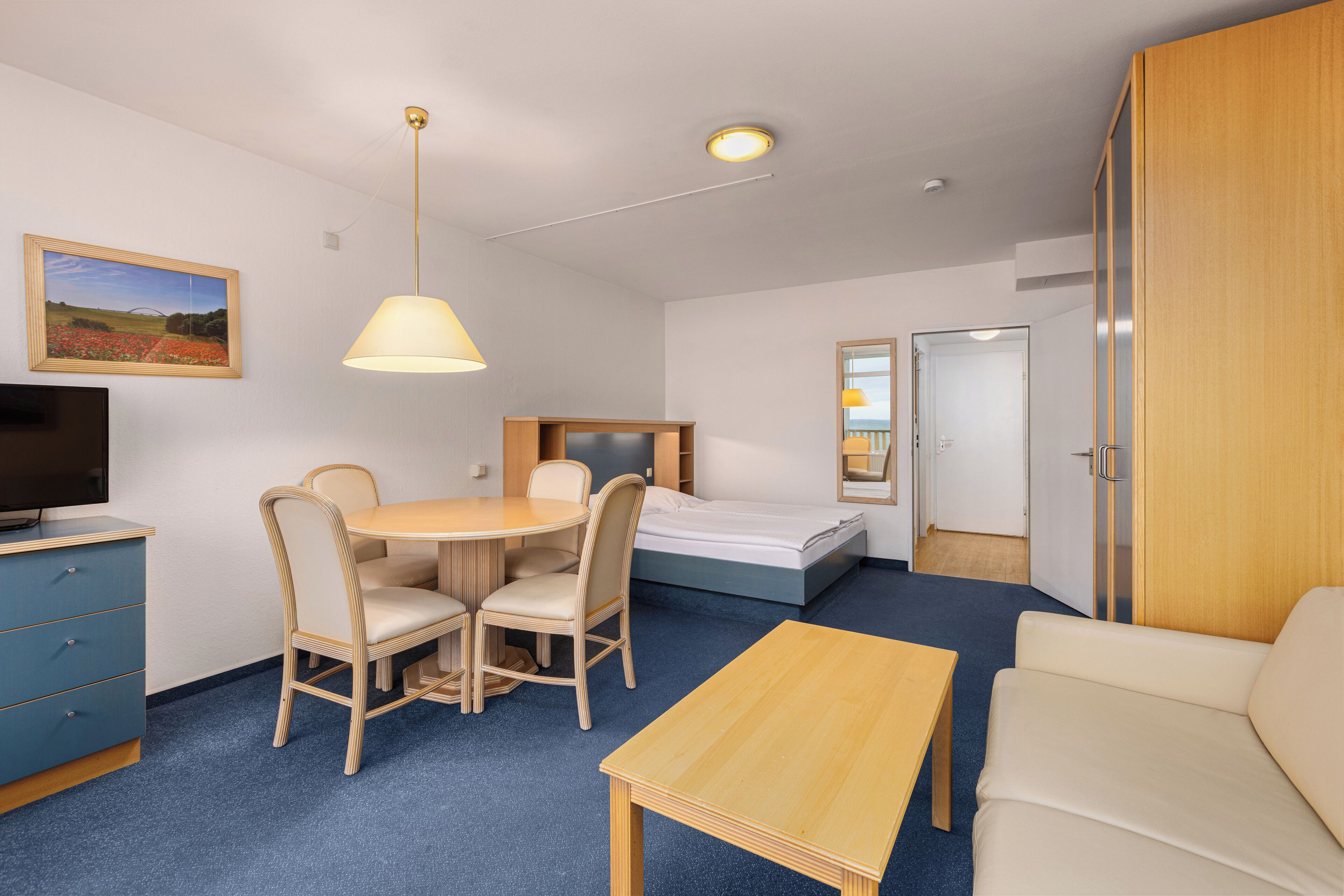 A hotel room with a bed, a desk with chairs, a TV, a lamp, and a wardrobe.