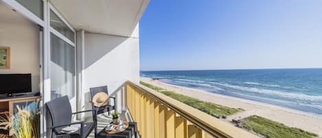 Een balkon met uitzicht op het strand, een tafel met stoelen en een televisie aan de linkerkant.