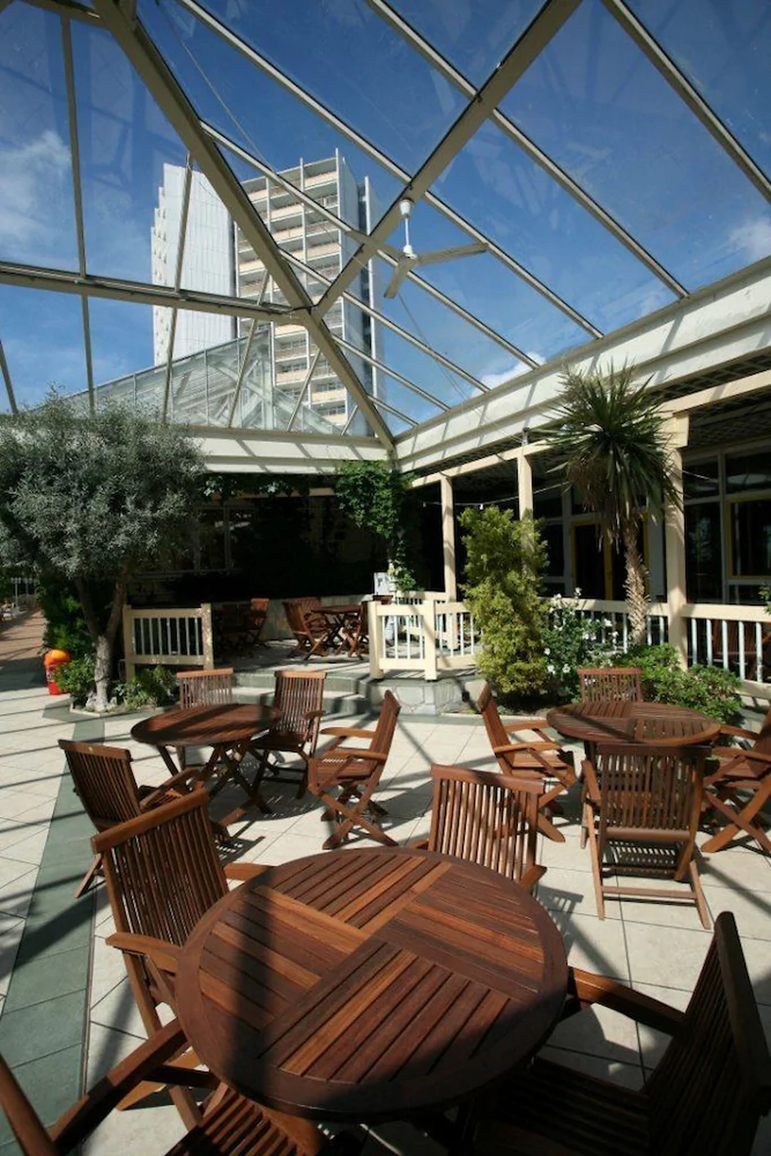 An outdoor seating area with wooden furniture under a glass roof, surrounded by greenery and a view of a building in the background.