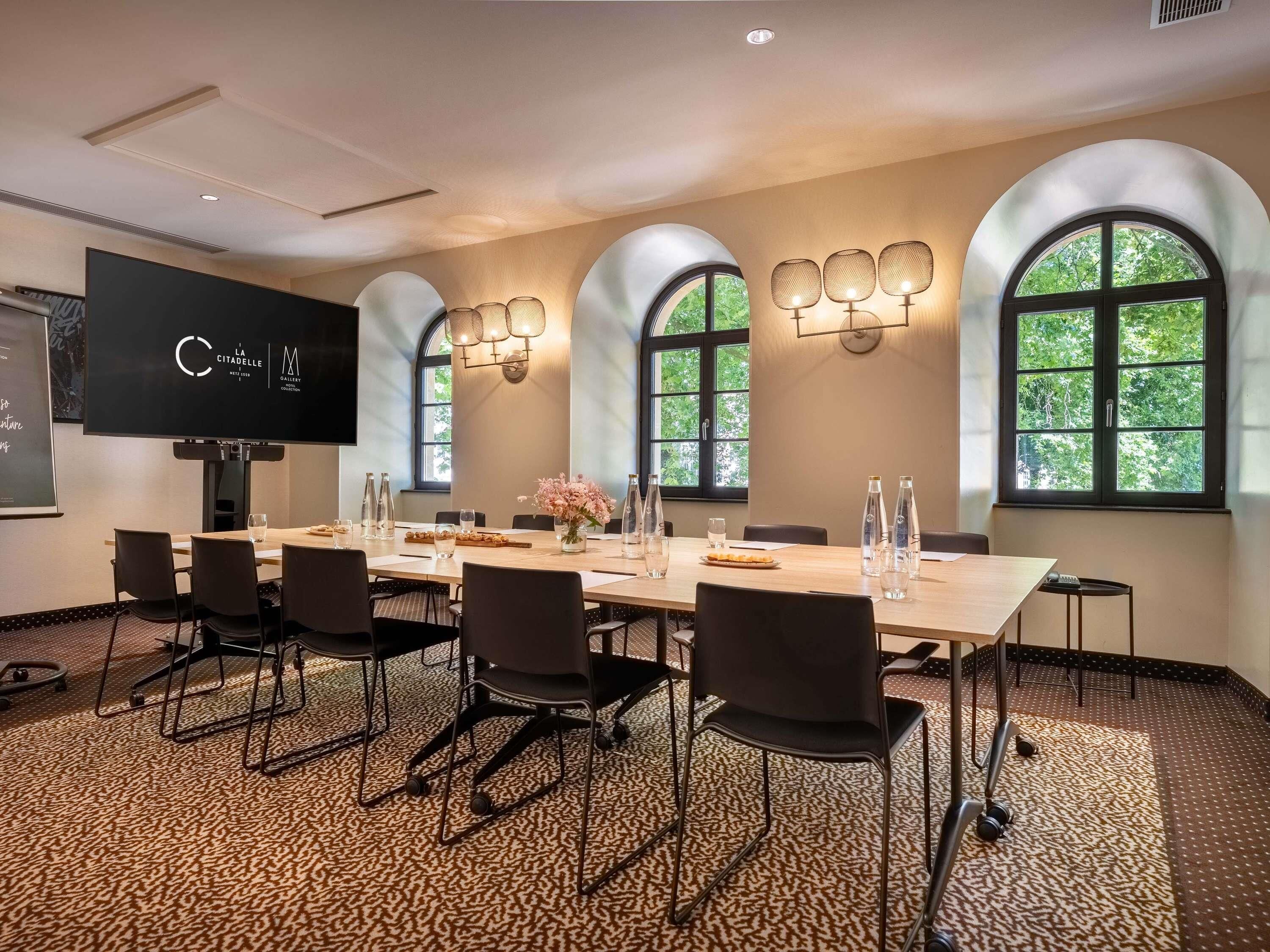A conference room with a long table, chairs, a projector screen, and arched windows with natural light.