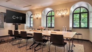 A conference room with a long table, chairs, a projector screen, and arched windows with natural light.