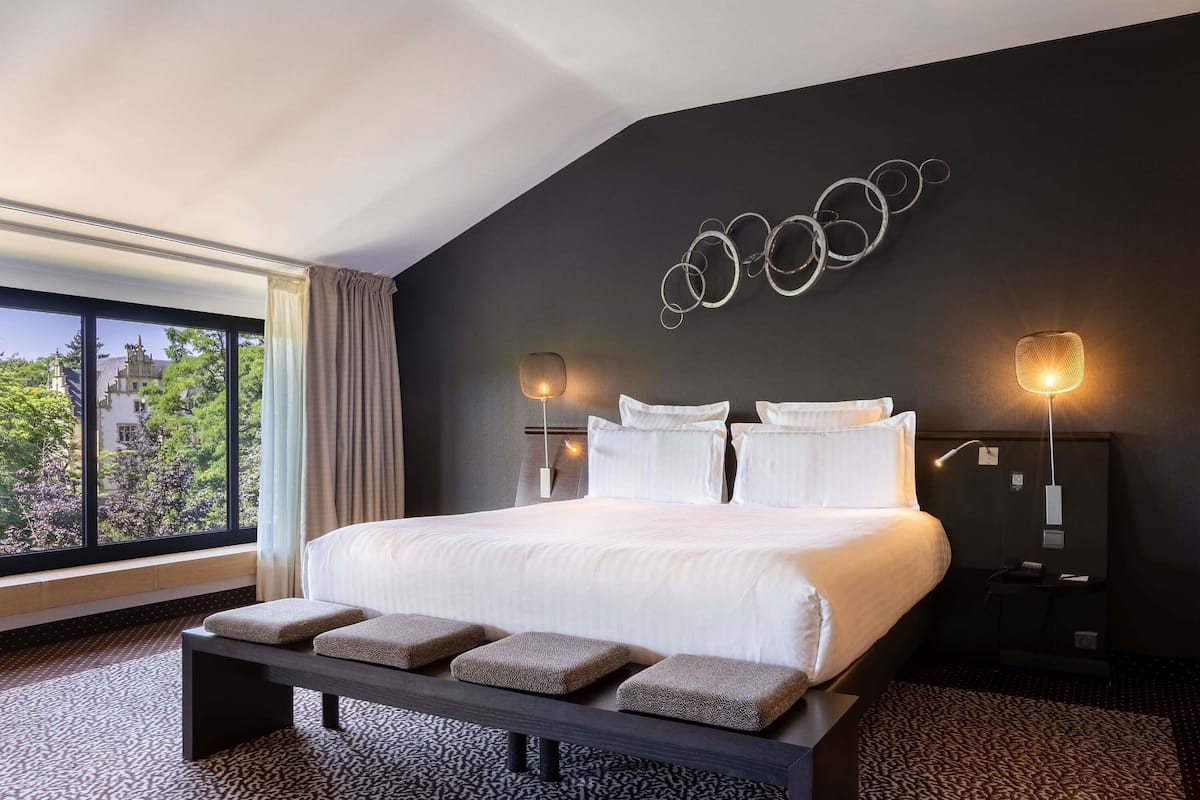 A modern hotel room with a large bed, a bench, two bedside tables, and a view of trees and buildings through a large window.