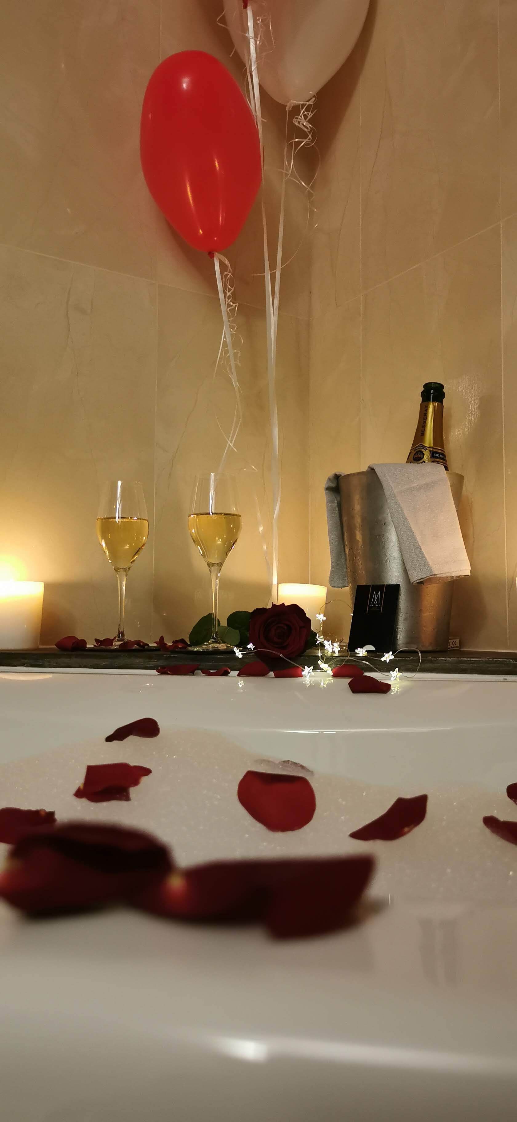 A bathtub with rose petals, two champagne flutes, a bottle of champagne, and a red balloon.