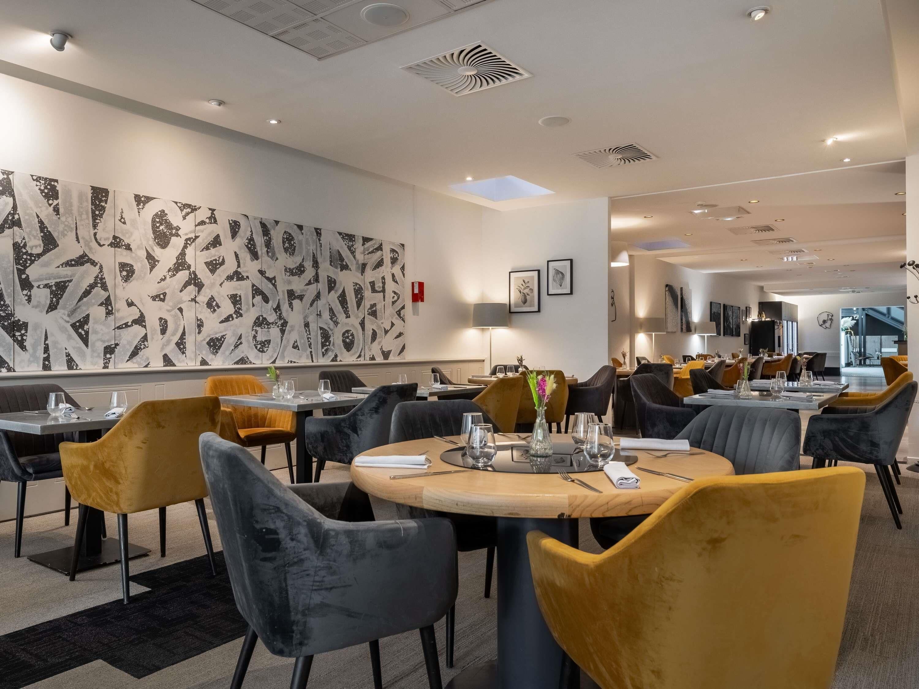 A modern dining area with round tables, black and yellow chairs, and a large abstract wall art piece.