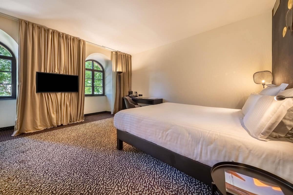 A hotel room with a bed, a desk, a chair, a TV, and a window with curtains.