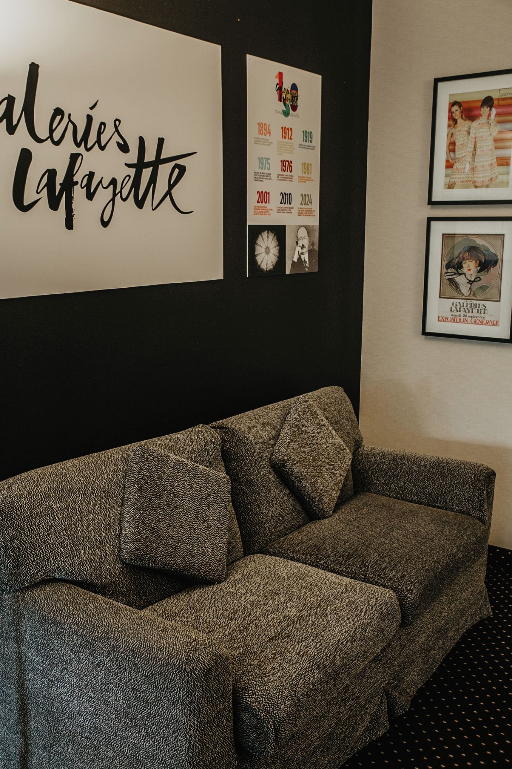 Ein Zimmer mit einer gemusterten Couch, einer Wand mit dem Schild 'Galeries Lafayette' und gerahmten Bildern.