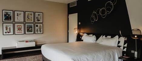 A modern hotel room with a large bed, a nightstand, a lamp, and a wall with framed posters.